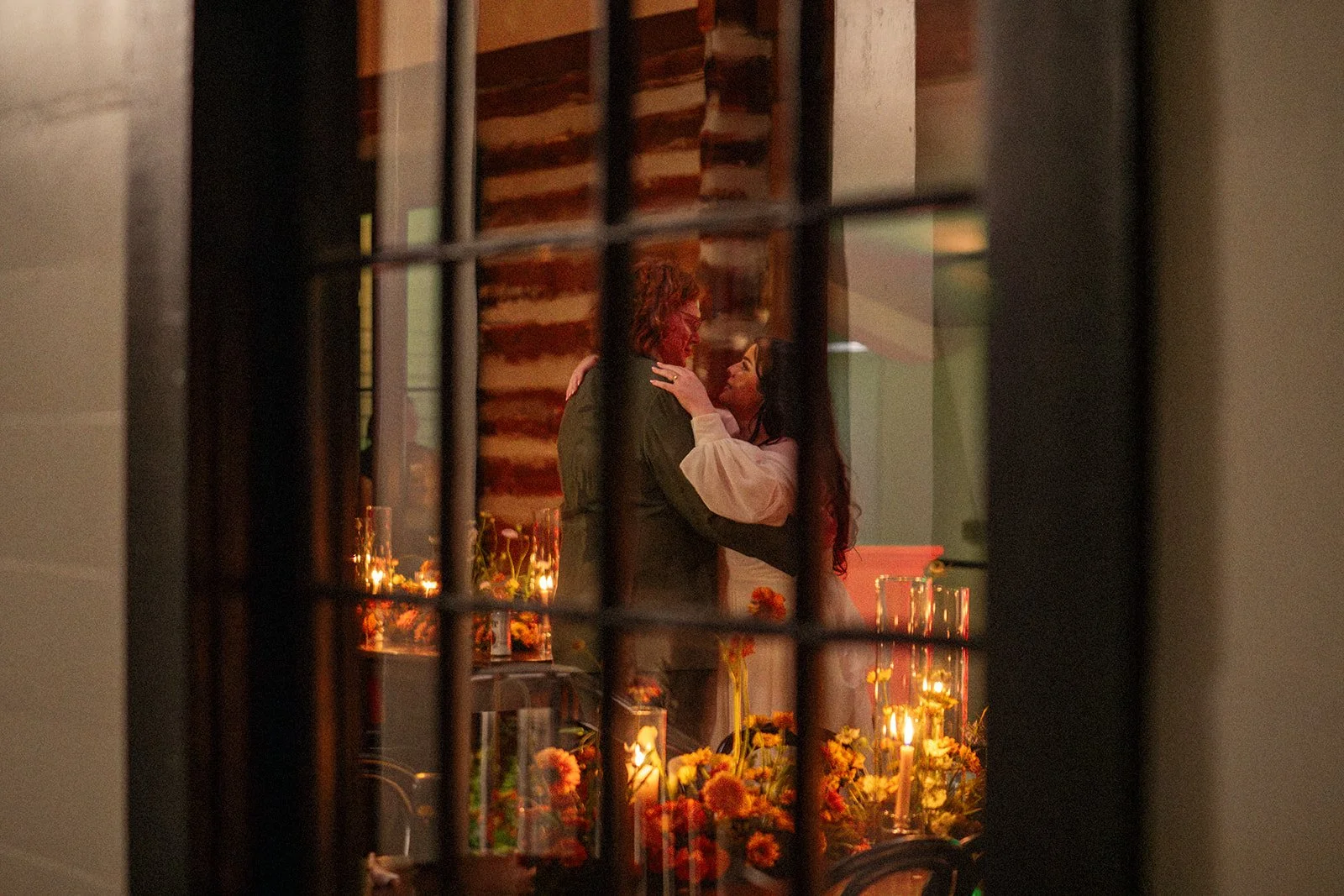 A couple dancing closely at a romantic dinner table decorated with flowers and candles, viewed through window bars.