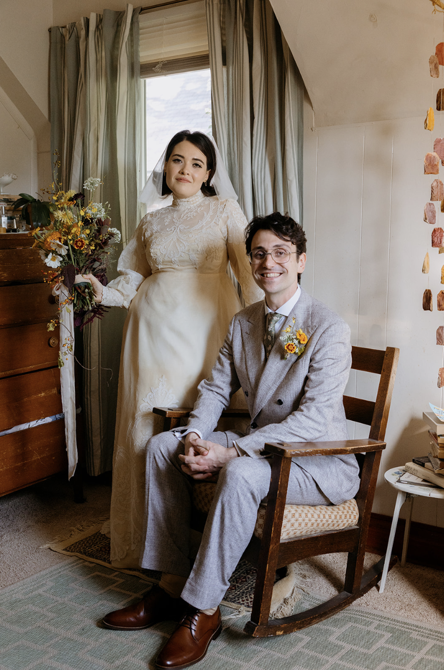 A woman in a vintage wedding dress holding a bouquet stands next to a man in a gray tuxedo with a sunflower boutonniere, sitting on a wooden rocking chair in a cozy room with a window, bookshelf, and colorful wall decorations.