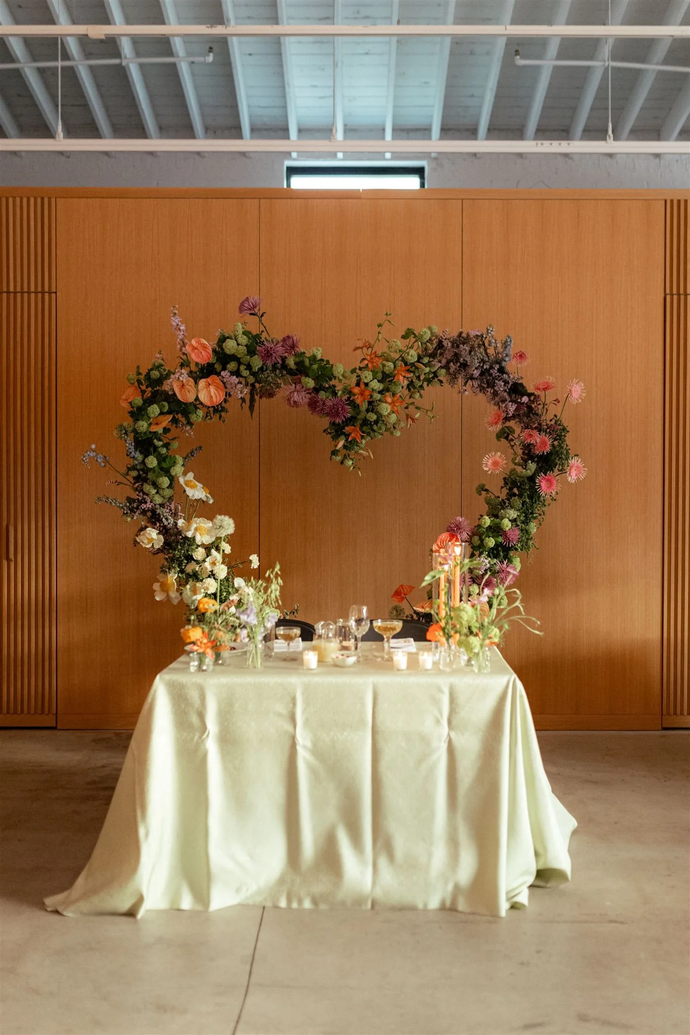 A table decorated with a large floral heart-shaped arch and candles in a spacious room with a wooden wall and a concrete floor.