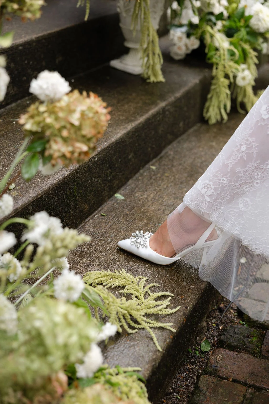 Close-up of a bride's foot in a white high heel with decorative embellishments, stepping onto a wet stone surface, surrounded by white and pink flowers and green foliage.