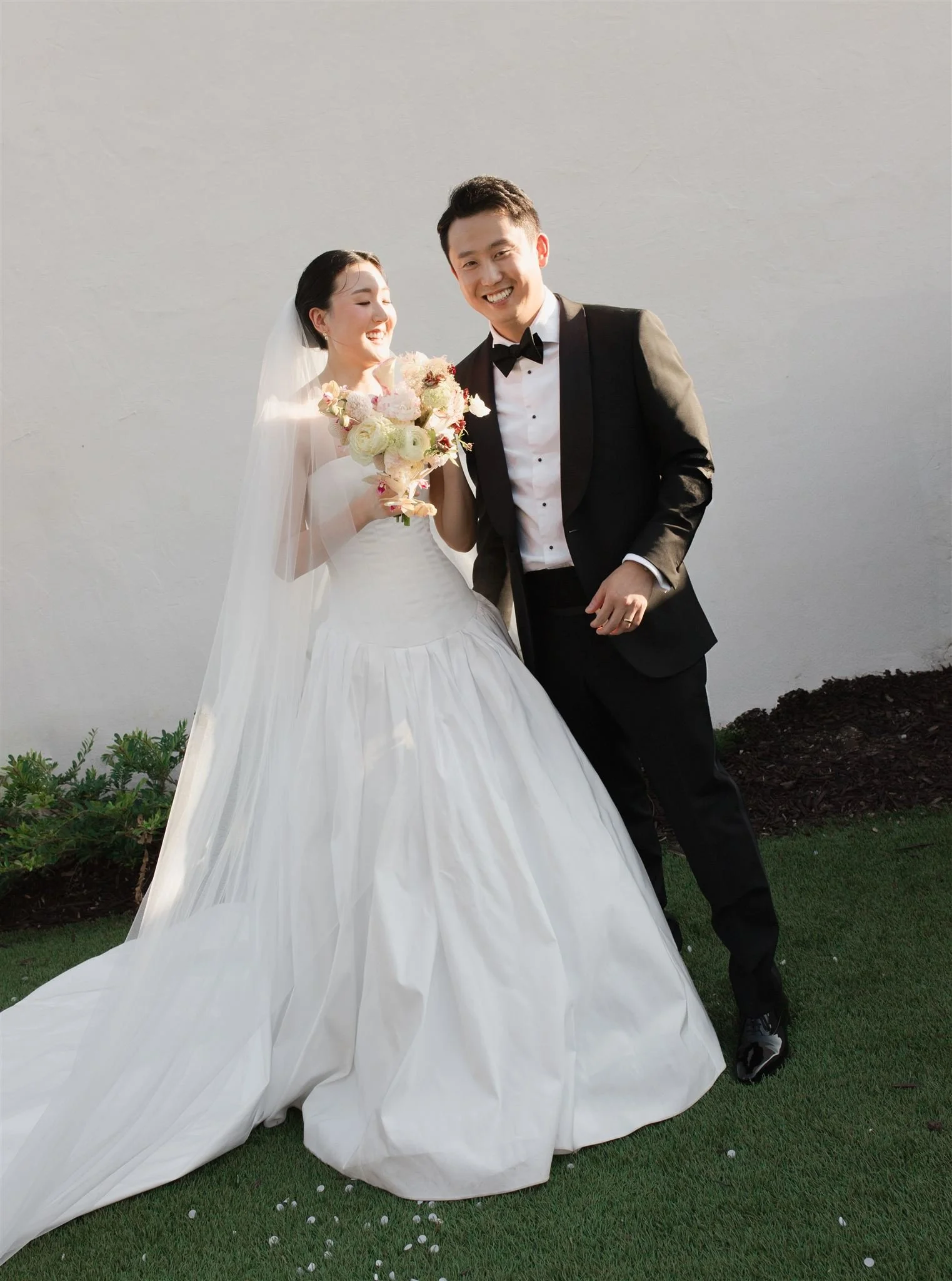 A newlywed couple dressed in wedding attire, standing on grass against a plain wall. The bride is holding a bouquet, wearing a white wedding gown with a long veil, and the groom is in a black tuxedo with a white shirt and black bow tie. They are smil