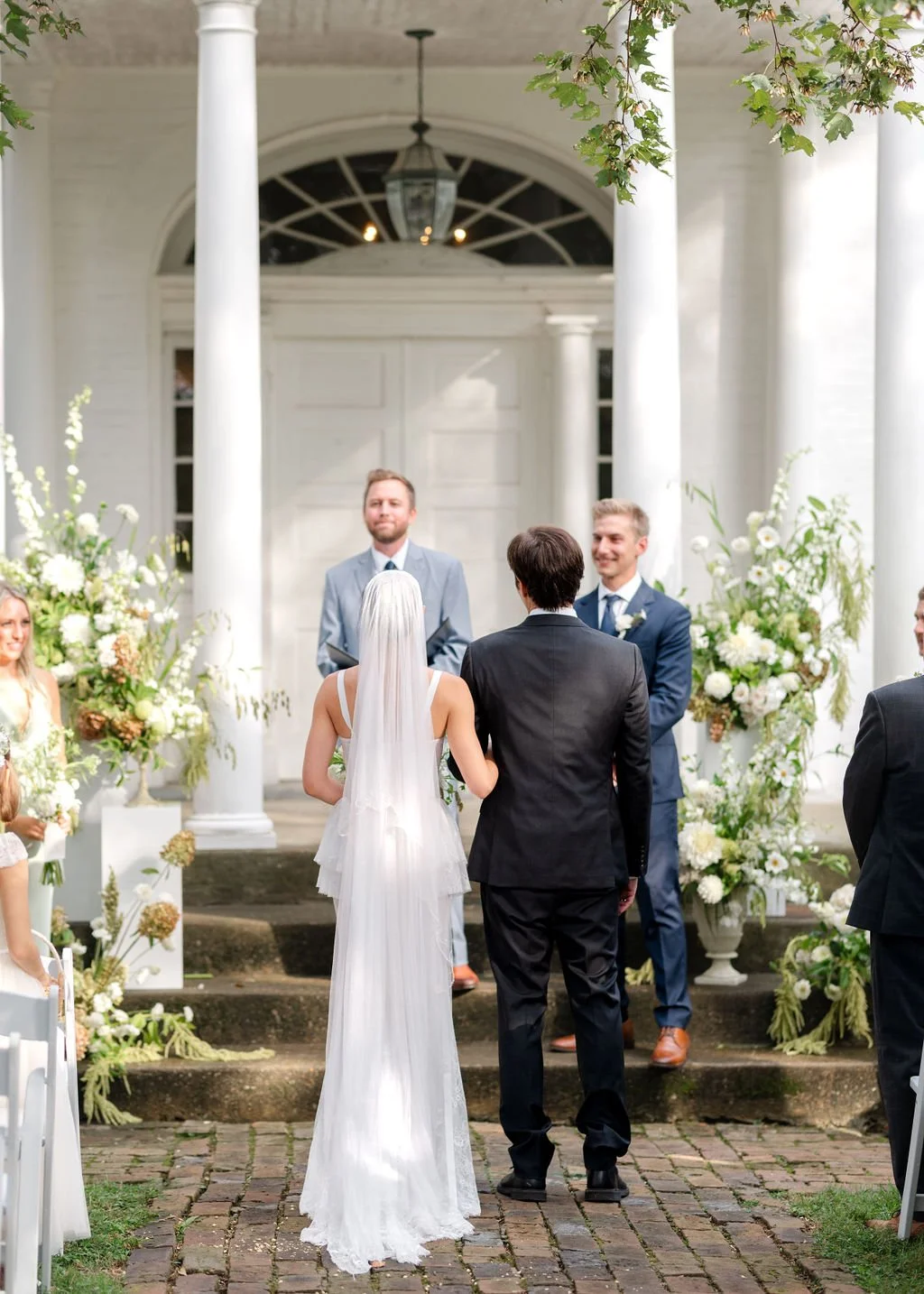 A couple getting married during an outdoor wedding ceremony on a brick pathway, surrounded by floral arrangements, with the officiant and guests in attendance.