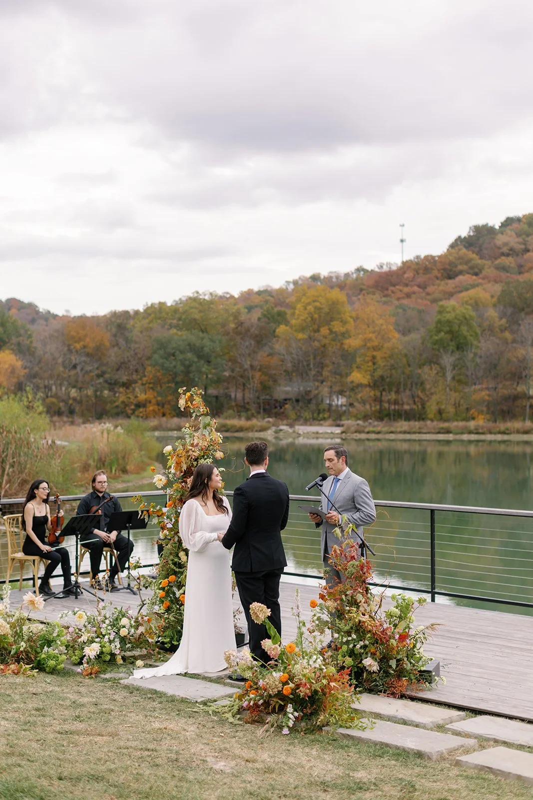 Bride and groom standing together during outdoor wedding ceremony beside a lake, with officiant reading from a tablet, surrounded by floral arrangements. Musicians sitting nearby with autumn foliage in the background.