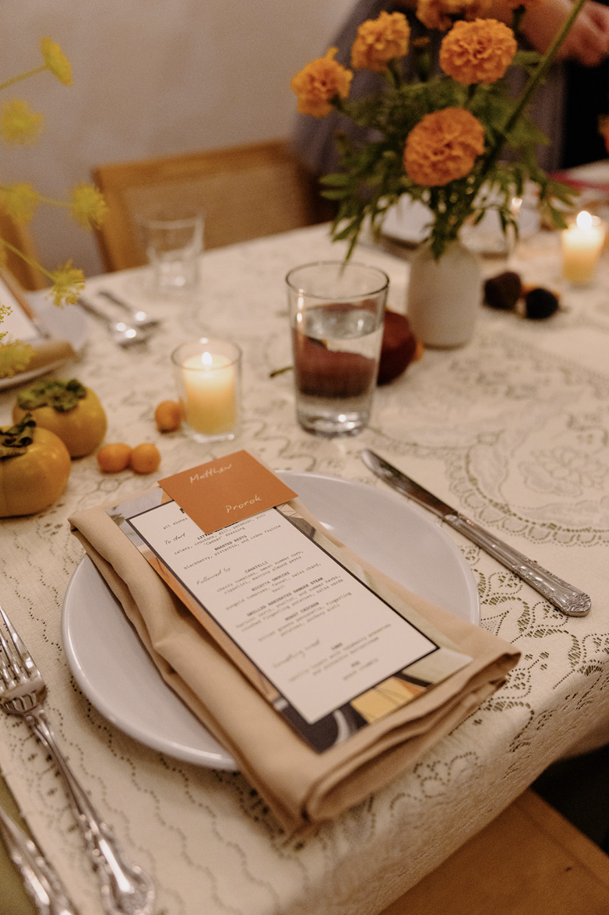 A close-up of a dining table set for a meal with a white lace tablecloth, a white plate with a folded napkin and menu card, silverware, a glass of water, small candles, yellow persimmon fruits, and a vase with orange flowers.