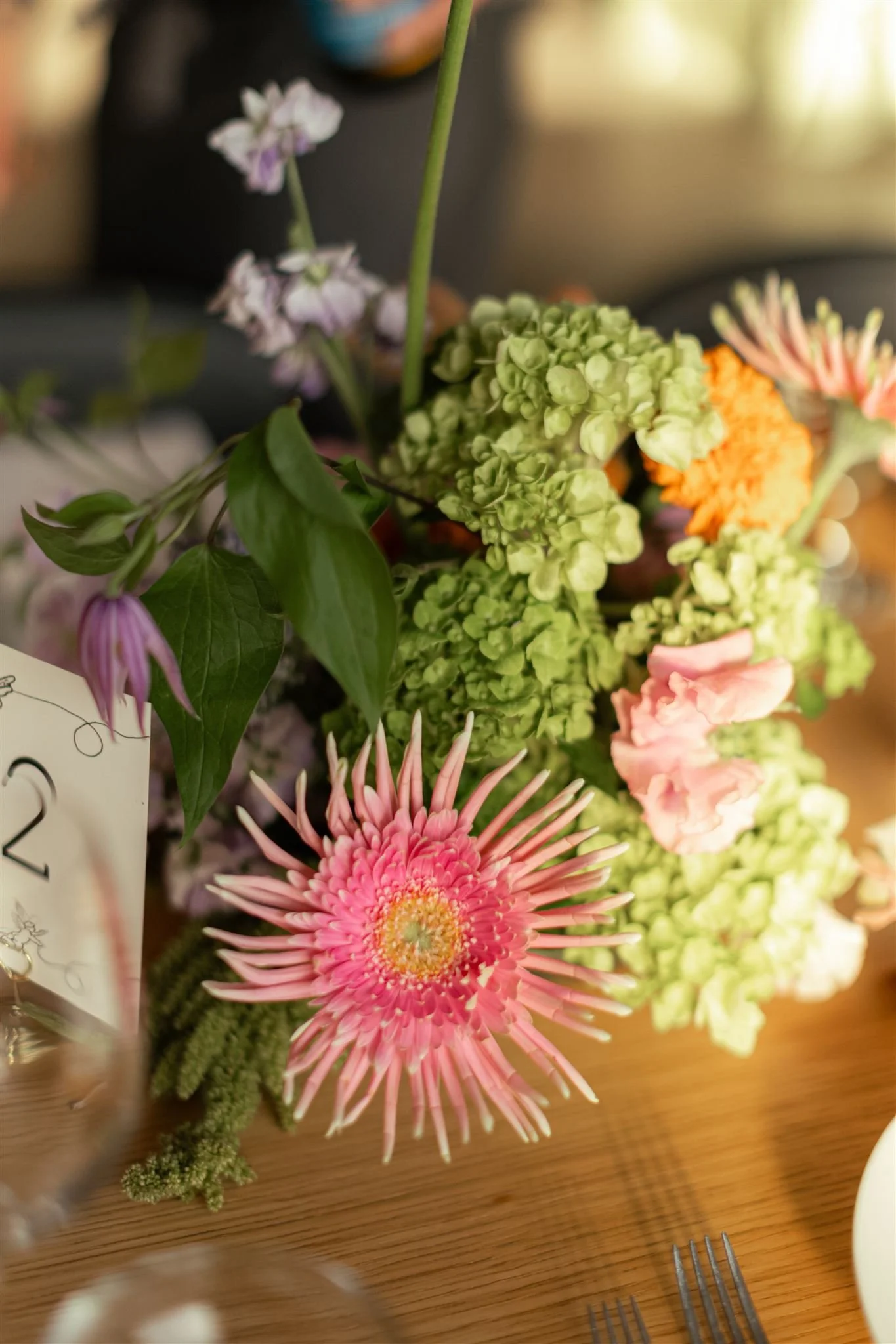 Close-up of a colorful flower arrangement with pink, orange, green, and purple flowers on a wooden table.