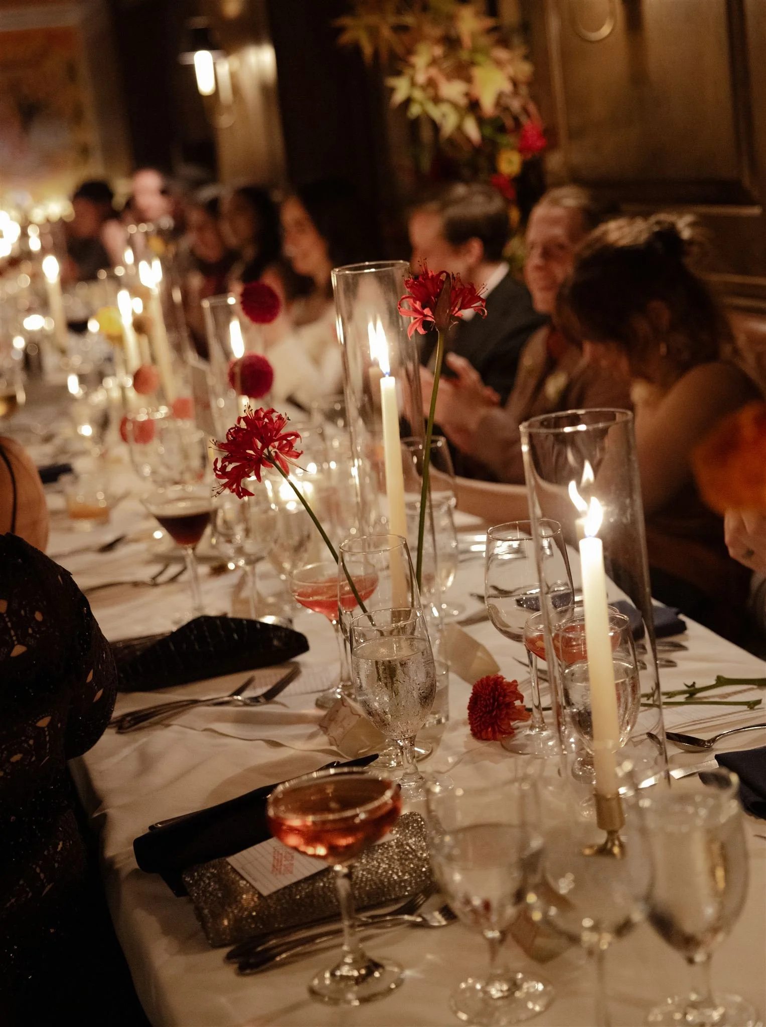 A formal dinner table decorated with red and pink flowers in glass vases, lit candles in tall glass holders, wine glasses filled with red and pink drinks, and elegant place settings. Guests are seated along the table, engaged in conversation, in a wa
