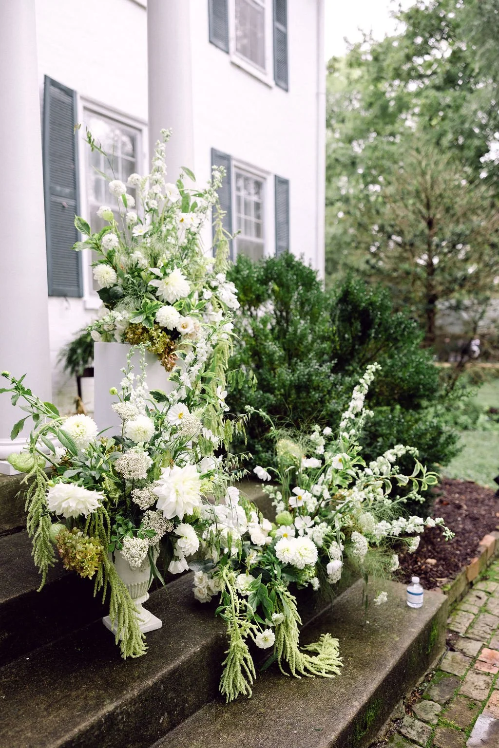 White floral arrangements on the steps of a house with white exterior walls and blue shutters, with greenery and trees in the background.