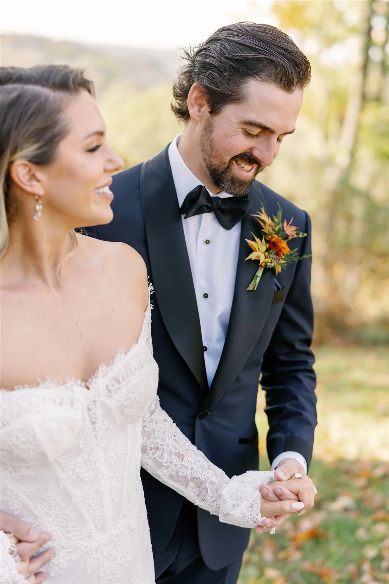 A bride and groom holding hands outdoors, smiling, with blurred trees and fall foliage in the background.