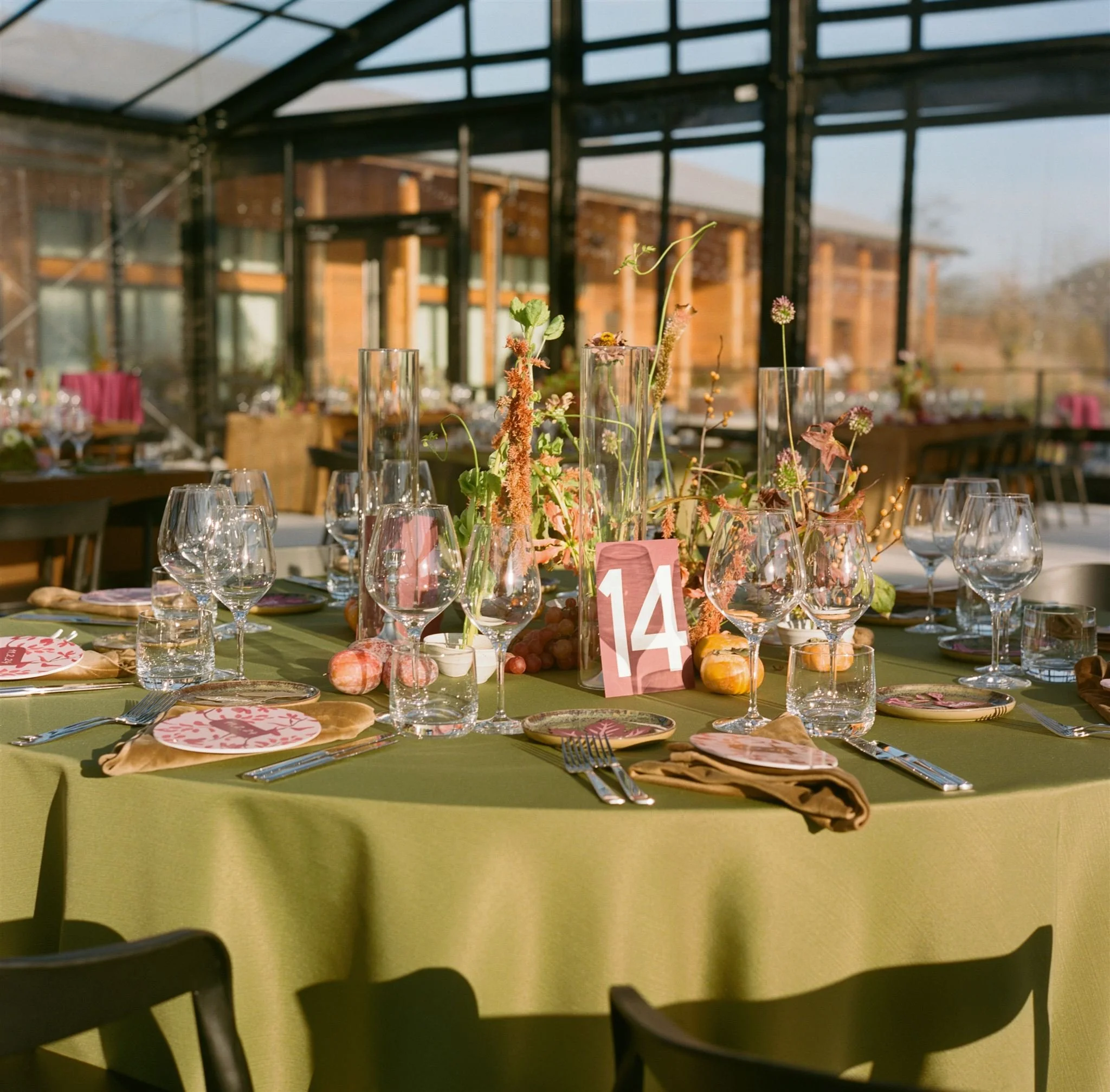 Round banquet table with a green tablecloth set with glassware, plates, and gold napkins, featuring a floral centerpiece with glasses and seasonal decorations. A pink table number card displaying '14' is in the middle. The background shows a glass-en