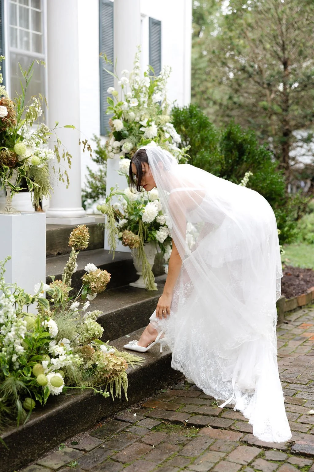 Bridal woman in a wedding dress adjusts her shoe on the steps outside a white house adorned with white and green floral arrangements.