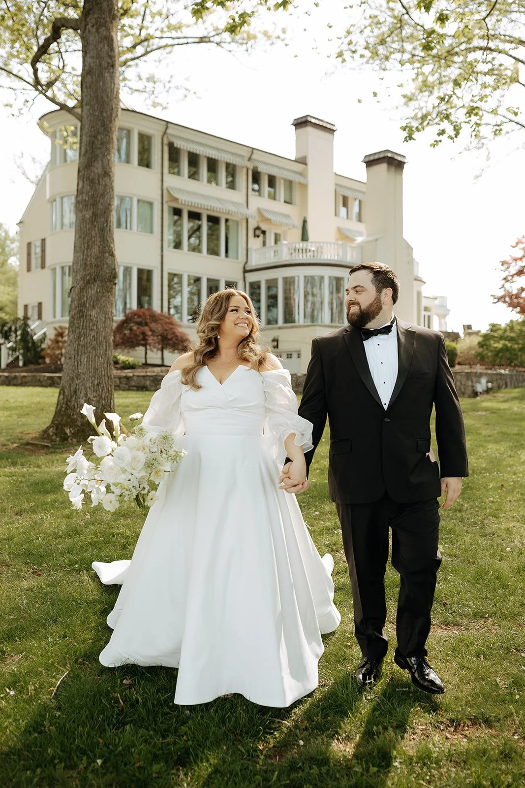 A bride and groom walking hand in hand on a grassy lawn, with a large mansion in the background. The bride is holding a bouquet of white flowers and wearing a white wedding gown, while the groom is in a black tuxedo.