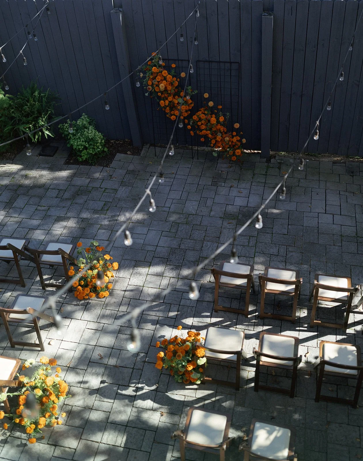 An outdoor patio area decorated with orange marigold flowers in pots and planters, surrounded by chairs, with string lights hanging overhead and shadows cast on the stone-paved ground.