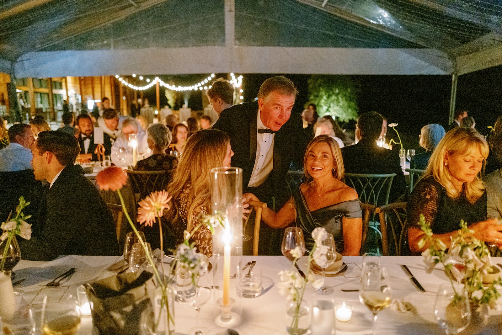 People at a formal dinner party, with a man in a tuxedo talking to two women sitting at a decorated table with flowers and candles, under a canopy at night, with other guests in the background.