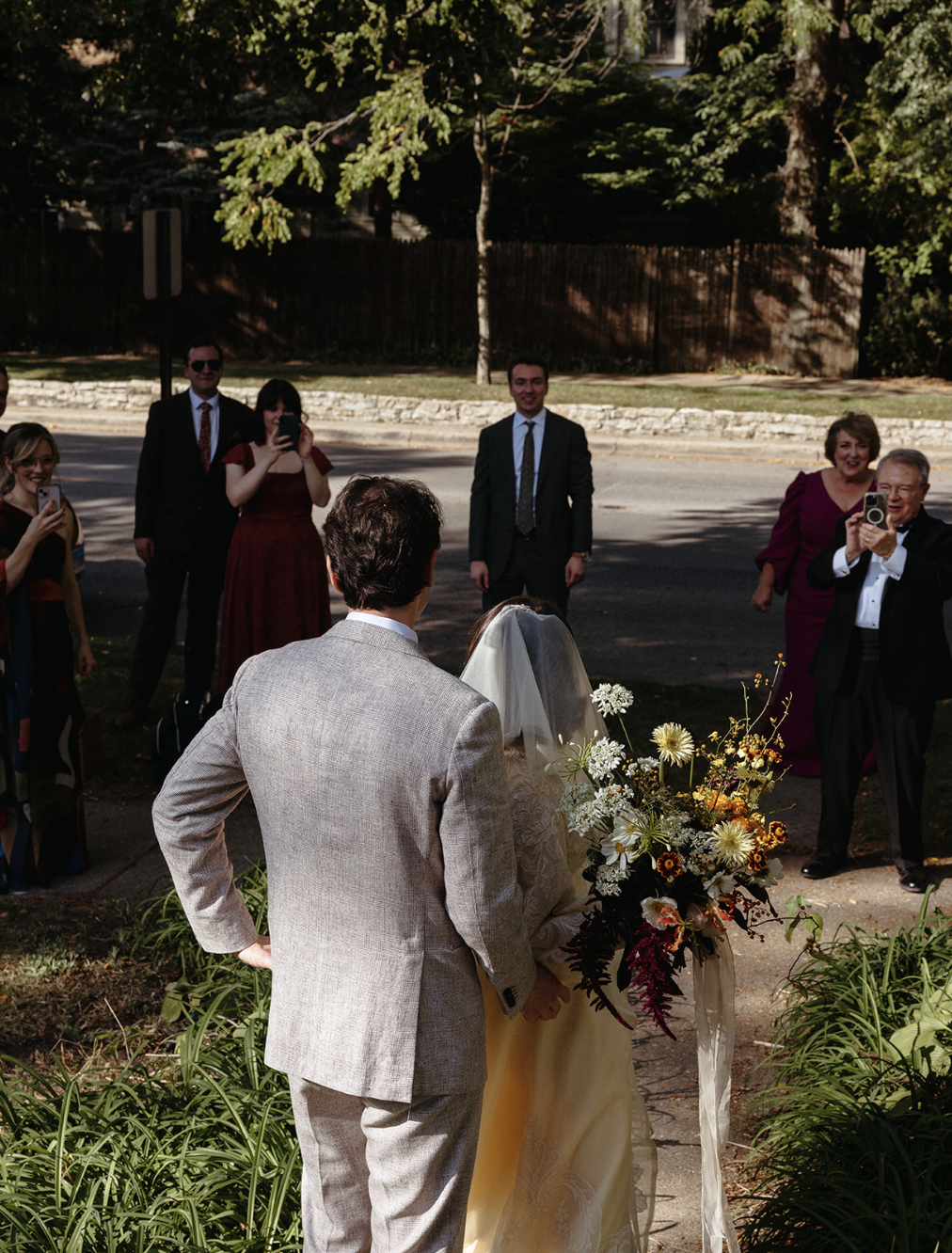 A bride and groom stand together outdoors, with the bride holding a bouquet of yellow, white, and dark red flowers. They are surrounded by guests taking photos during their wedding.