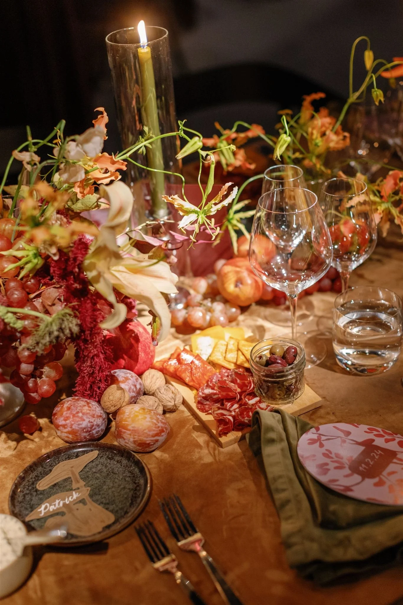 A festive table setting with a wooden table decorated with a floral arrangement, grapes, nuts, cheese, cured meats, and empty wine glasses. A tall candle is lit, and a paper plate with a group name and date is on a napkin.