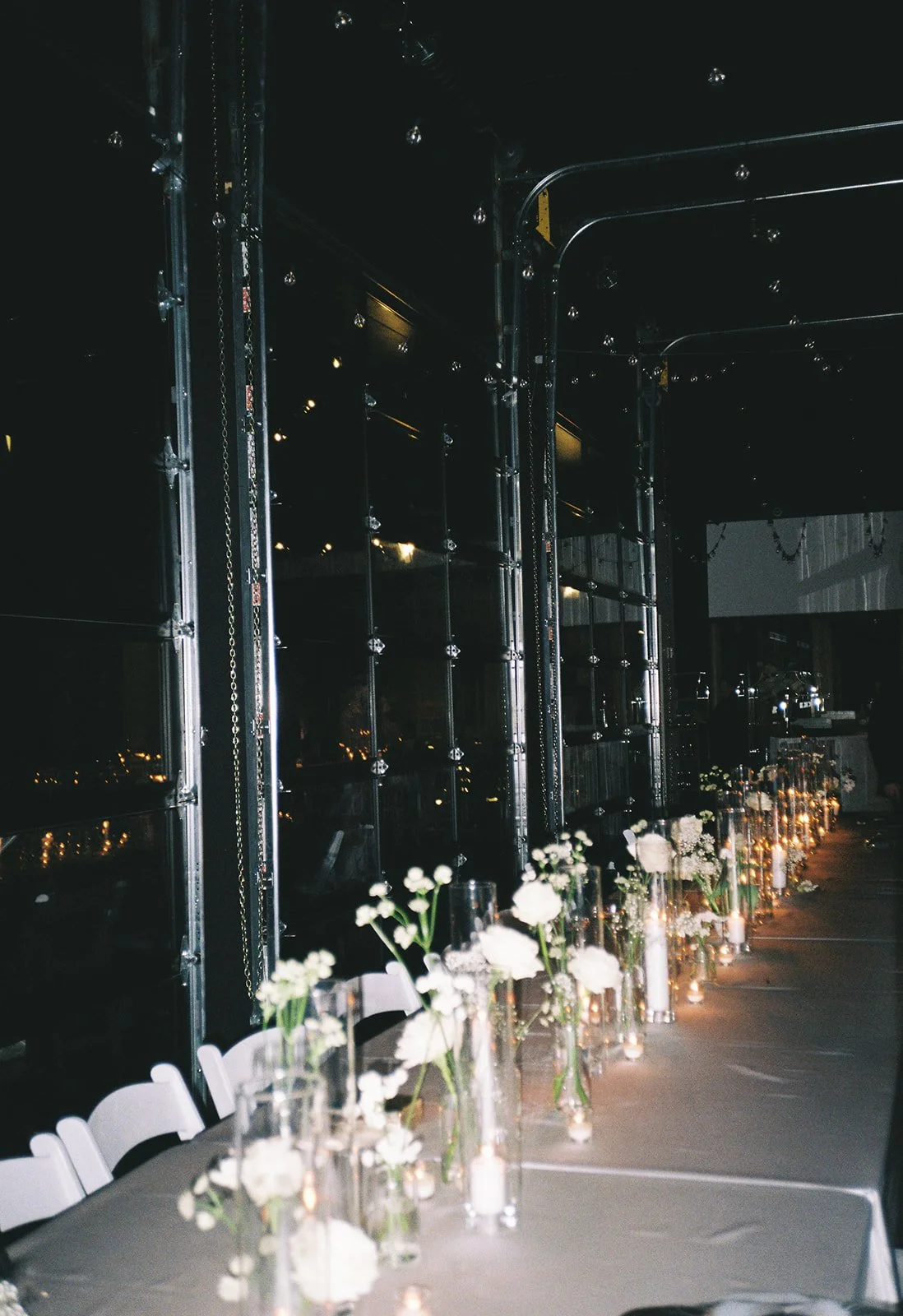 A long table decorated with white flowers and candles, set up for a nighttime event or celebration, with city lights visible through a glass wall