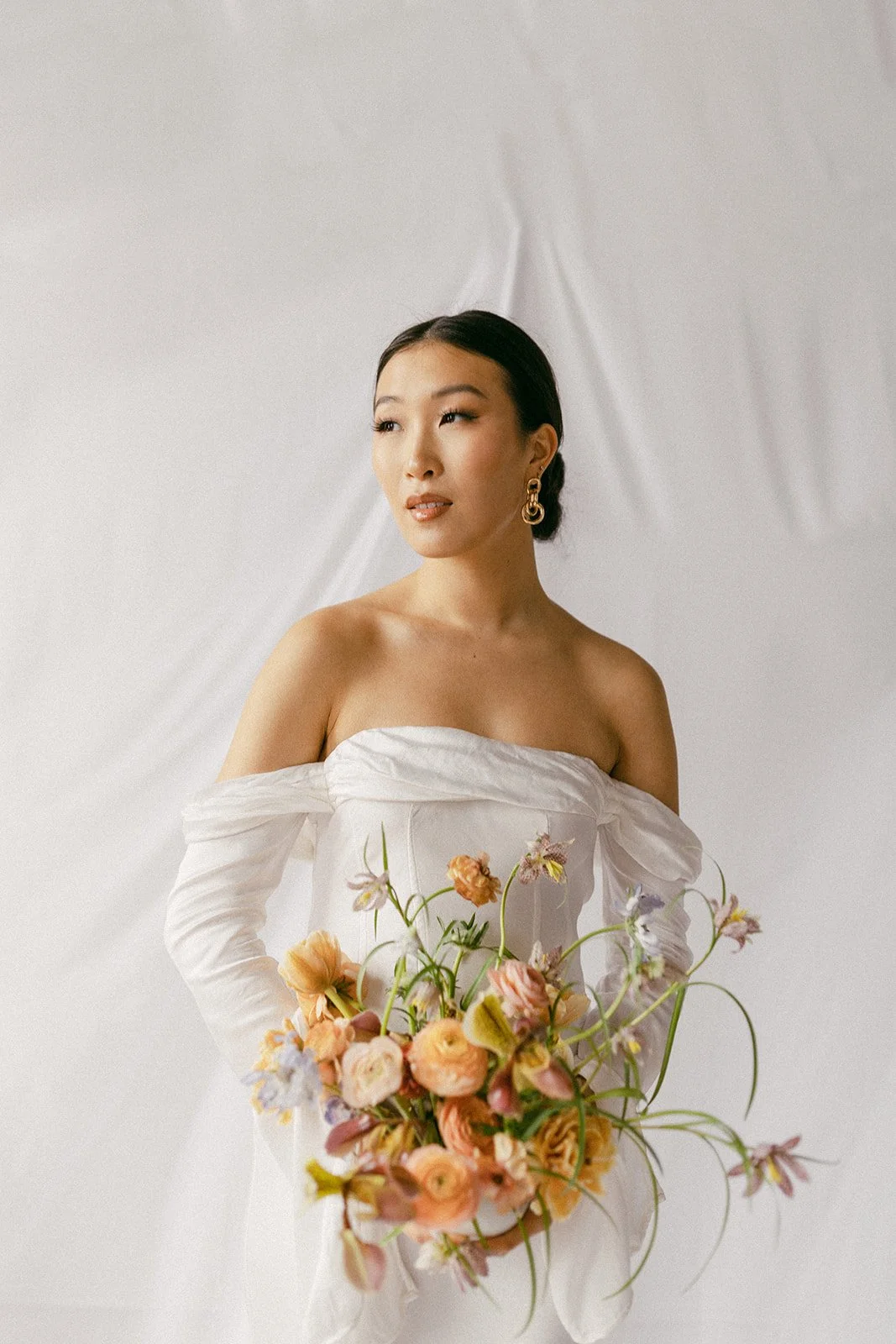 A woman wearing an off-shoulder white dress holding a bouquet of peach, pink, and purple flowers against a plain background.
