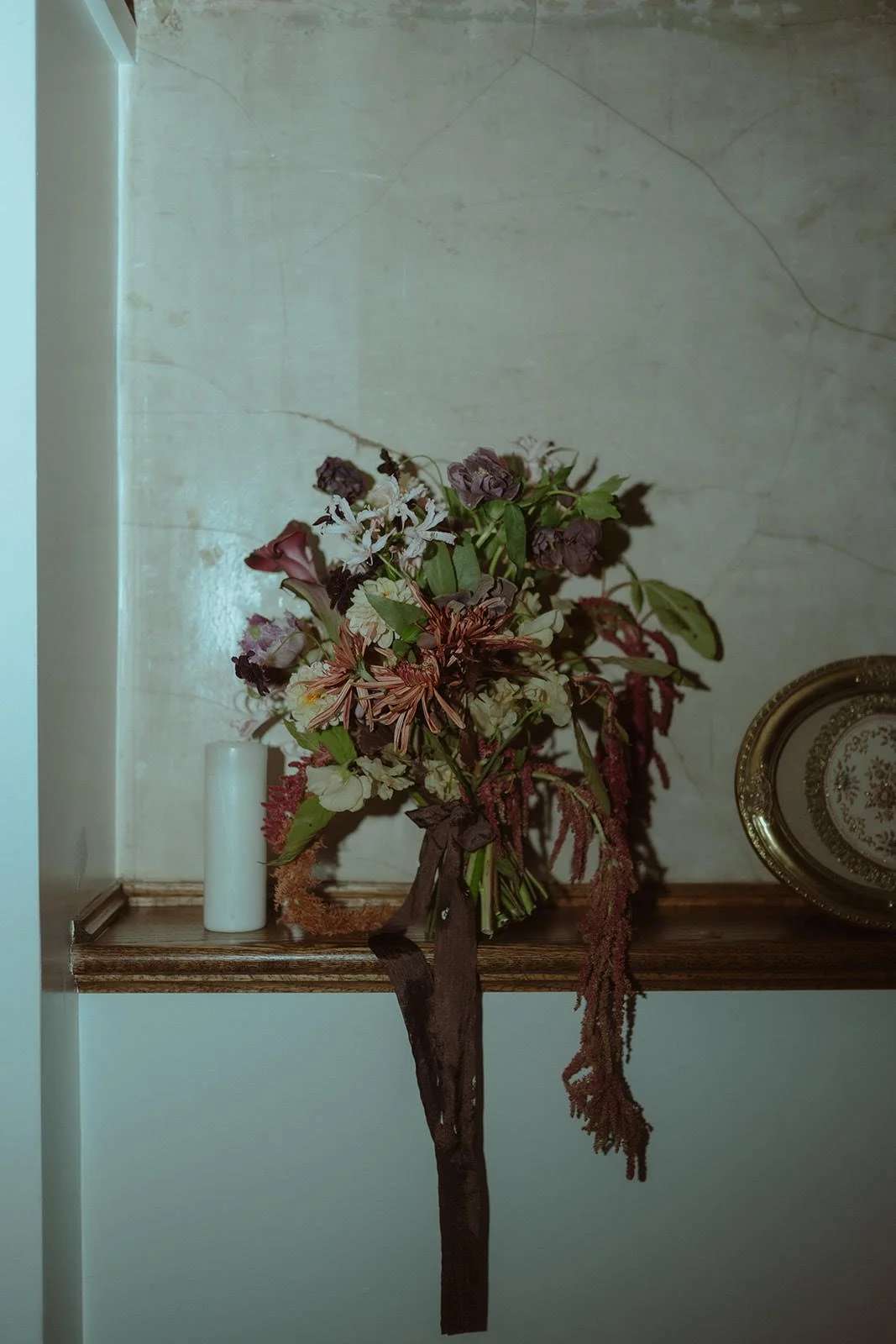 A floral arrangement in a vase with various flowers and greenery on a wooden shelf, with a textured wall and a decorative plate to the right.
