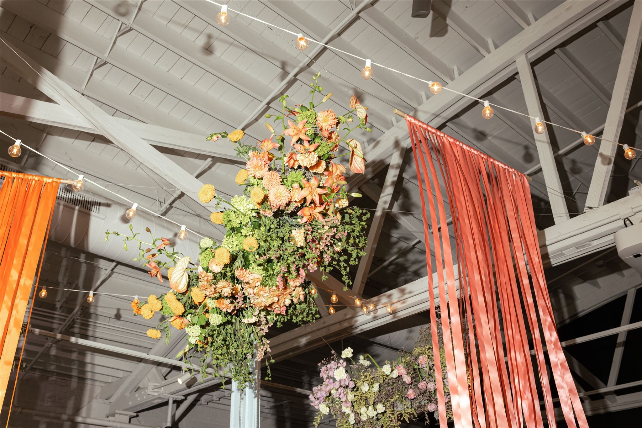 Colorful floral arrangement hanging from the ceiling with string lights and orange curtains in the background.