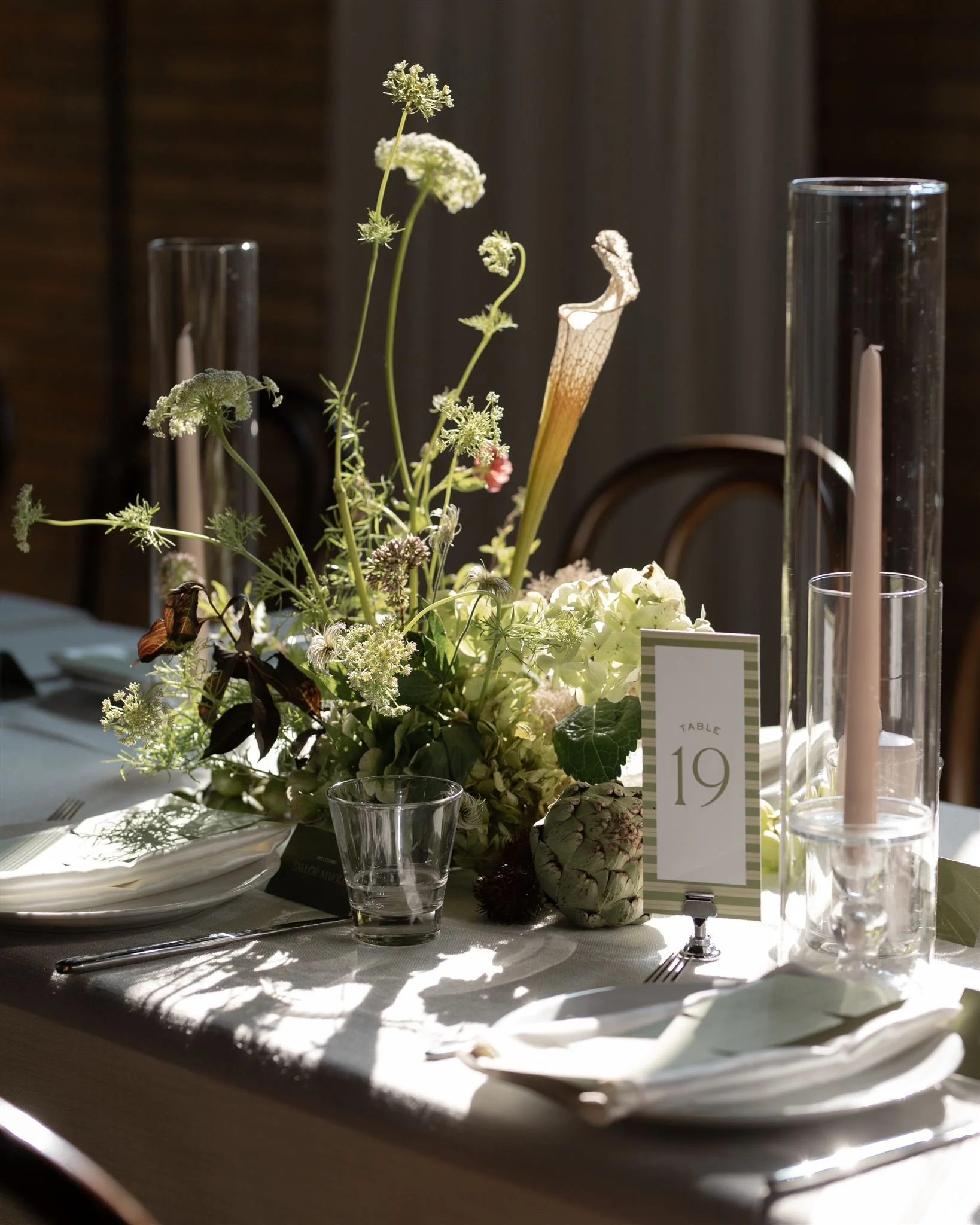 Elegant table centerpiece with white and green flowers, tall clear glass candle holders, a table number card reading 'Table 19', and place settings on a white tablecloth.