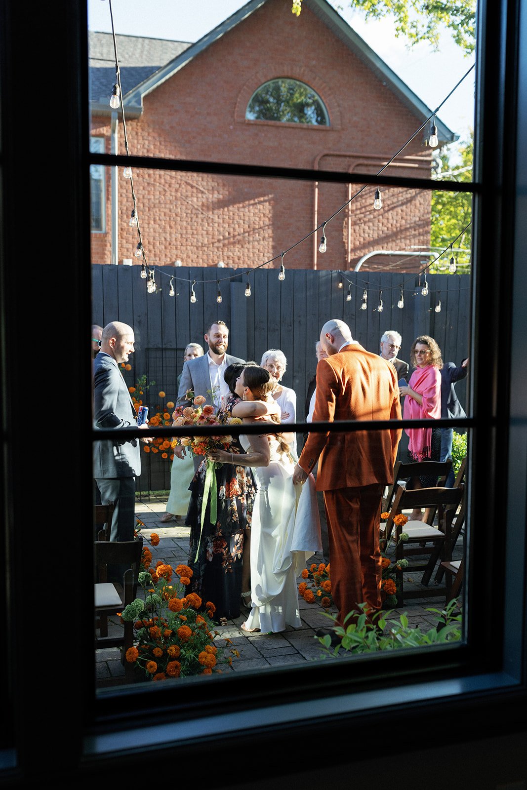 A wedding ceremony taking place outdoors in a backyard, viewed through a window. The bride and groom are embracing, surrounded by friends and family, with string lights overhead and orange flowers decorating the ground.