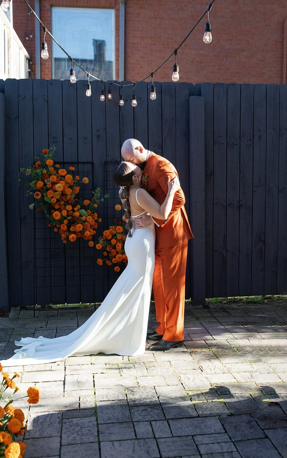 A couple sharing a romantic kiss at their wedding reception, with outdoor string lights above and orange flowers on a black fence behind them.