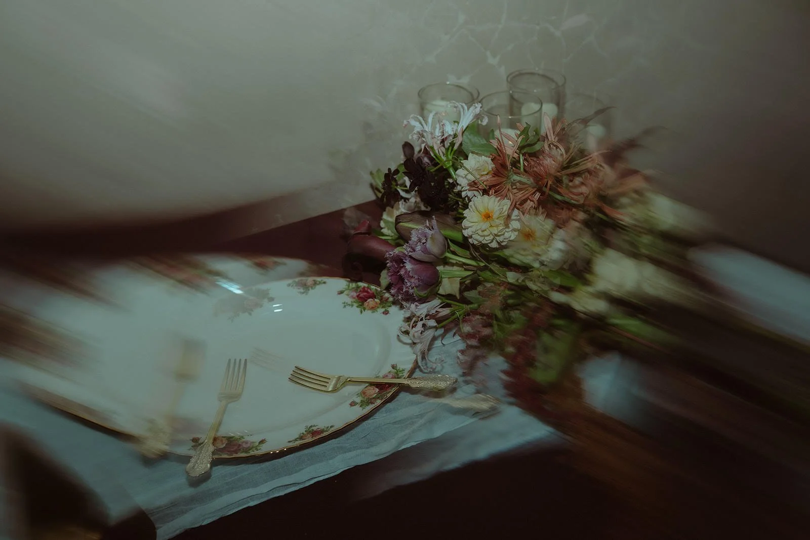 A dining table with floral tableware, a bouquet of flowers, and three glass candle holders in the background.