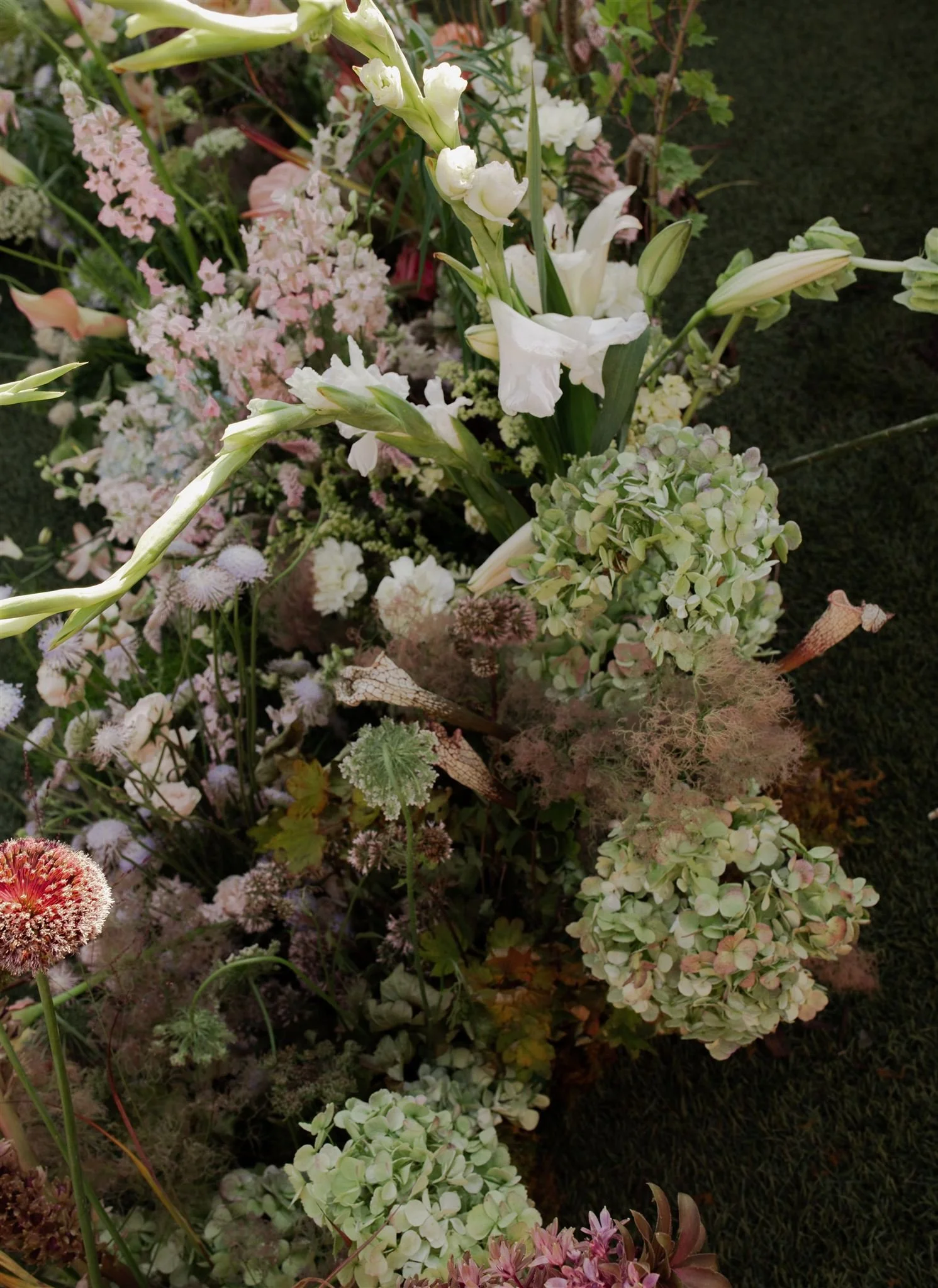 A close-up of a colorful flower arrangement featuring white lilies, pink and green hydrangeas, and various other small flowers and greenery.
