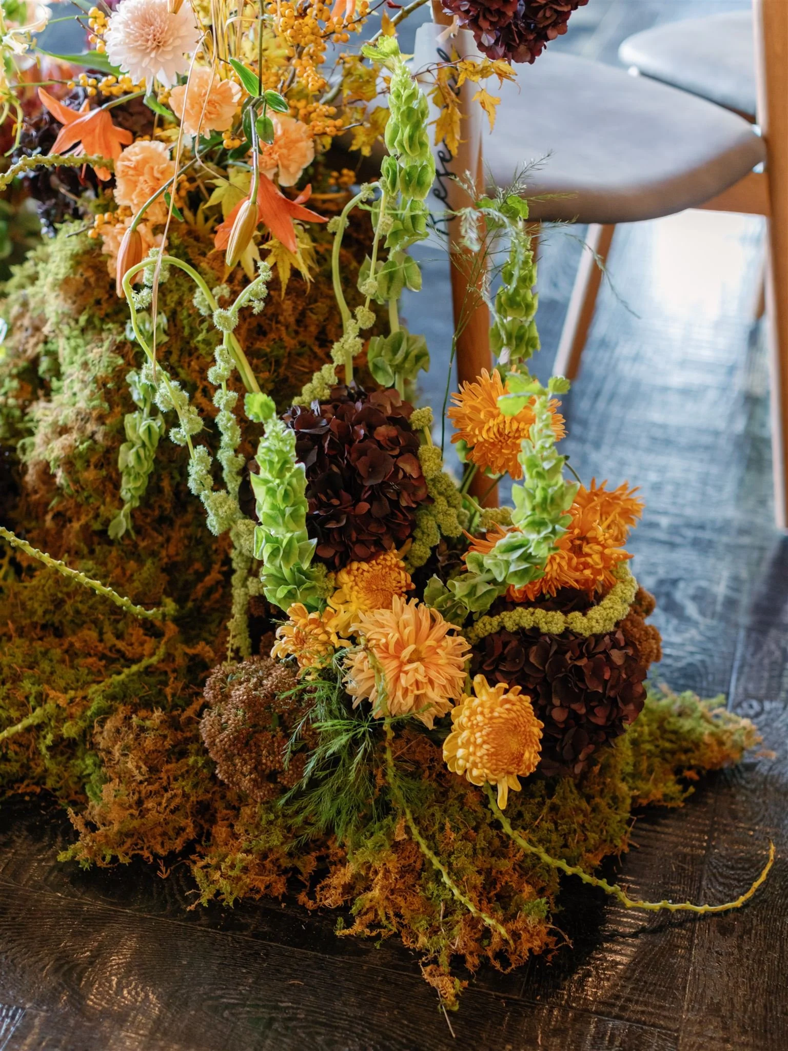 An arrangement of autumn flowers, including orange, yellow, and dark blooms, set among moss and green foliage on a dark wooden surface, with a chair in the background.
