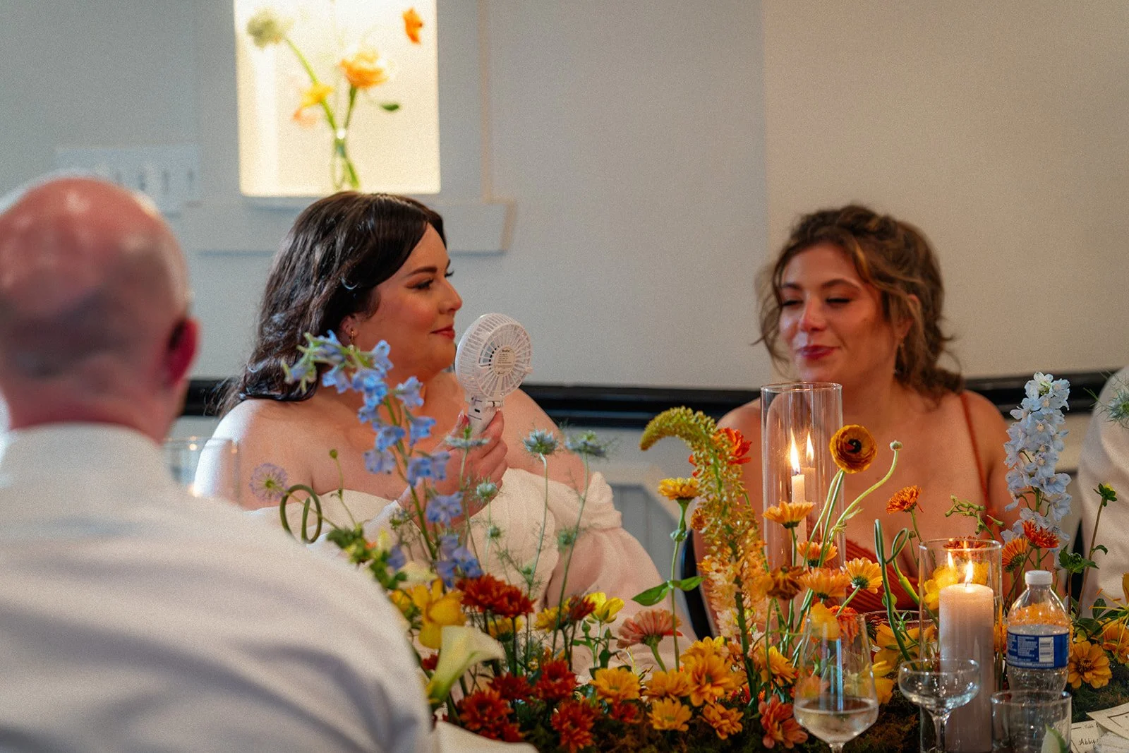 A woman sitting at a table with colorful flowers and candles, holding a small handheld fan, smiling while talking to another woman at a dining event.