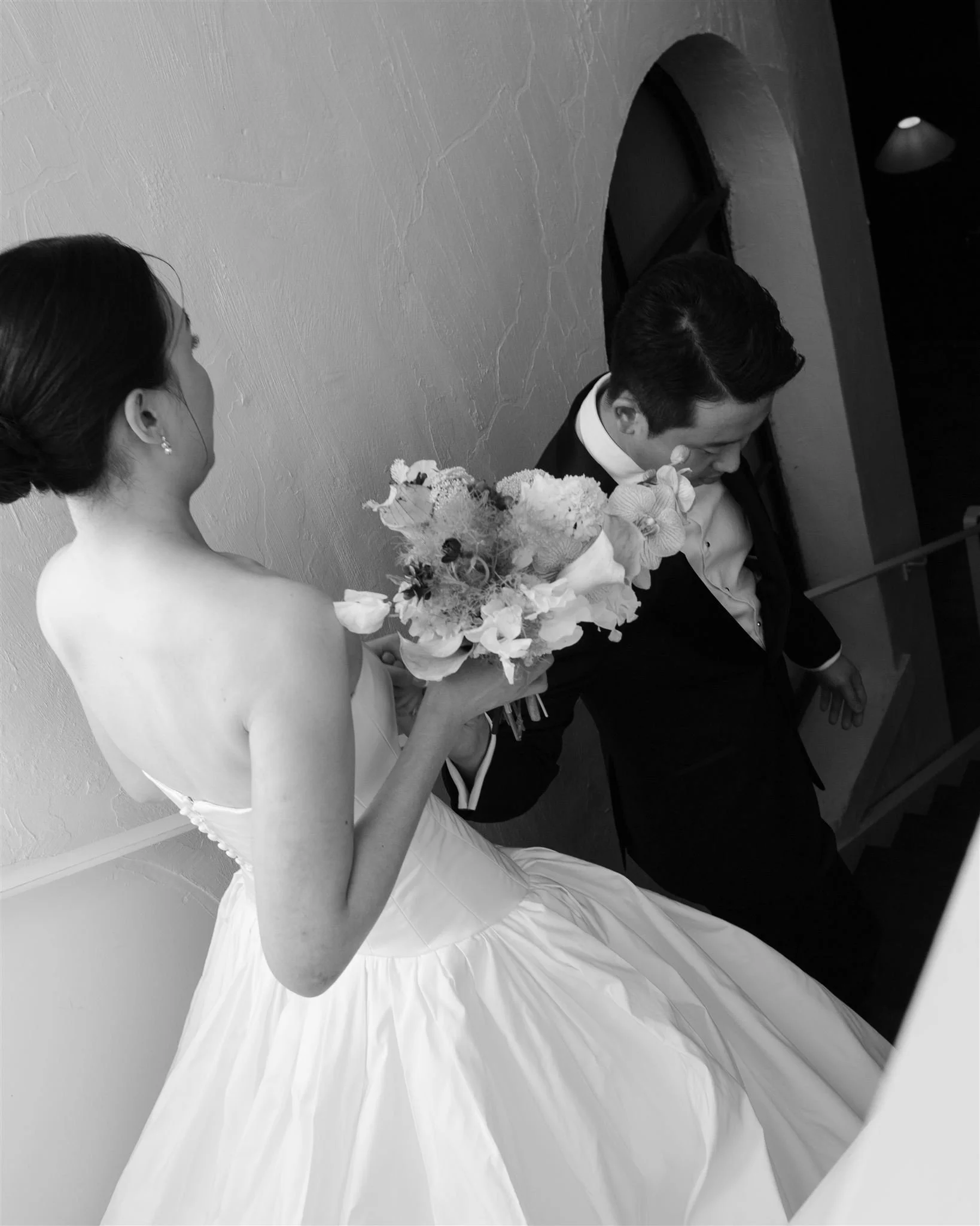 Black and white photo of a bride holding a bouquet while walking down the stairs with a groom. The bride is wearing a strapless wedding gown, and the groom is dressed in a tuxedo. The bride is smiling, and they are looking at each other.
