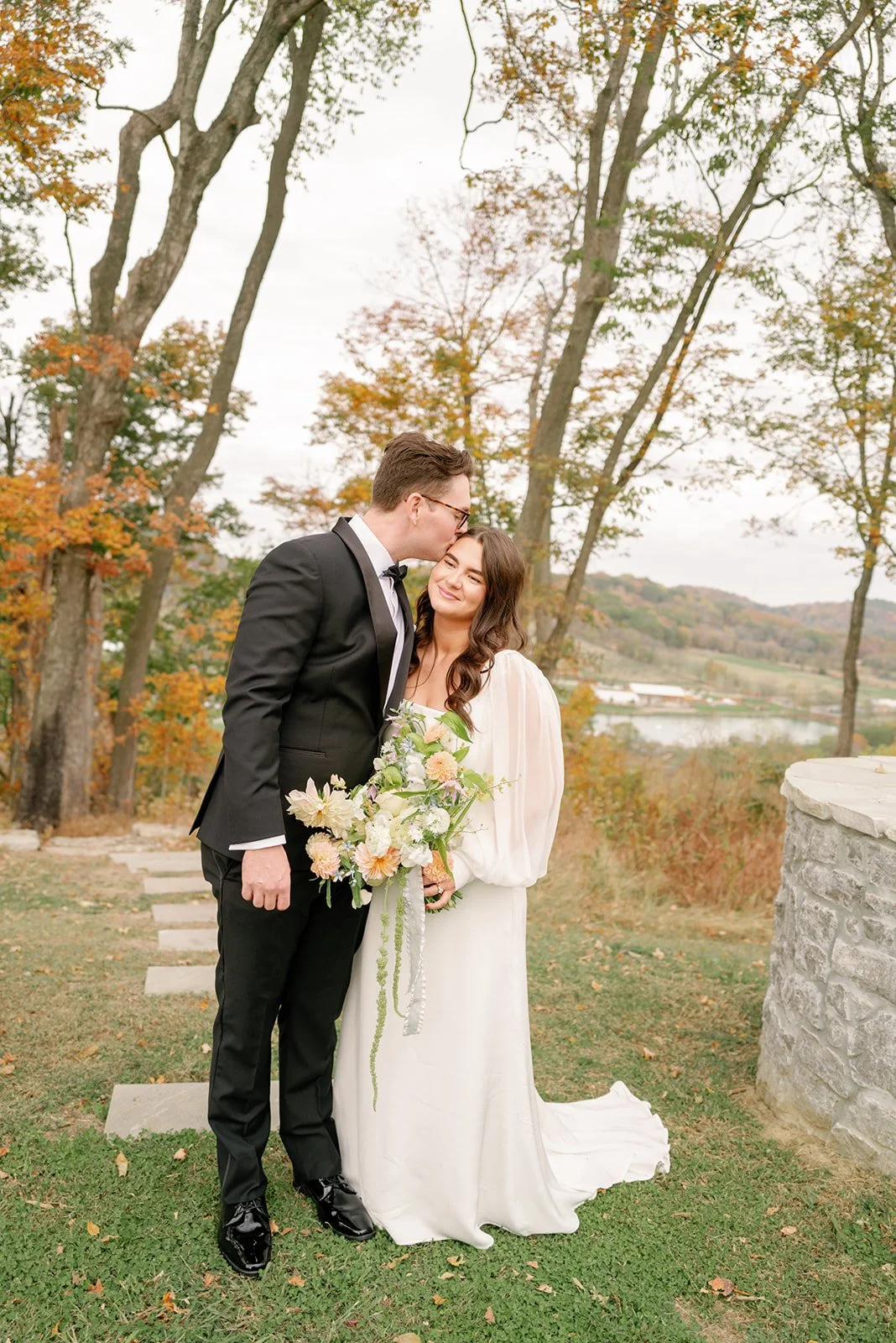 A bride and groom standing outdoors in an autumn setting, with the groom kissing the bride on her forehead. The bride holds a bouquet of flowers, and both are smiling. The background features trees with fall foliage, a stone structure, and a scenic l