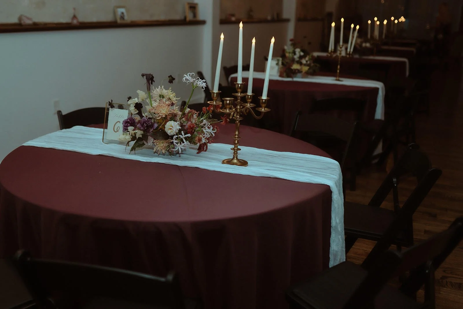 Round table decorated with a maroon tablecloth, white runner, floral centerpiece, and gold candelabra with lit candles. Additional tables in the background have similar decor and candlelight.