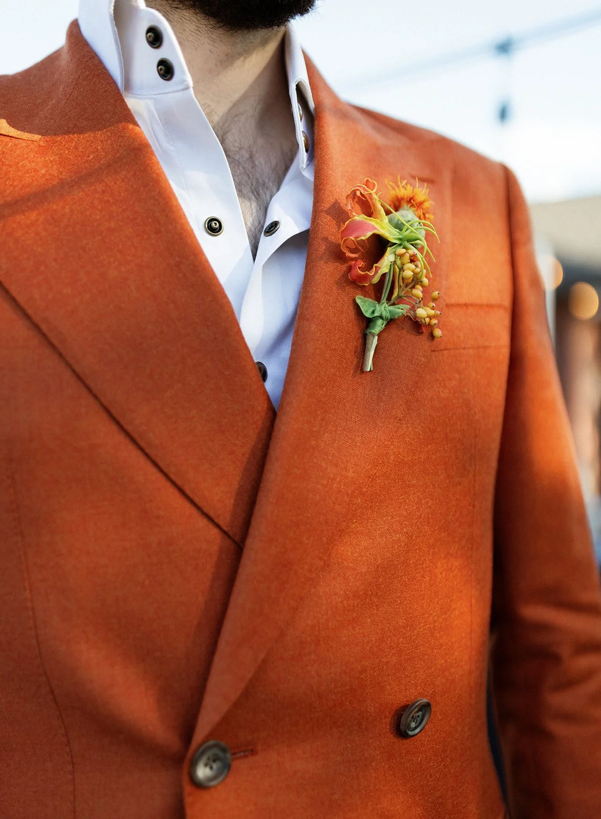 Close-up of a man wearing an orange blazer over a white shirt with button details. A floral boutonniere is attached to the blazer's lapel.