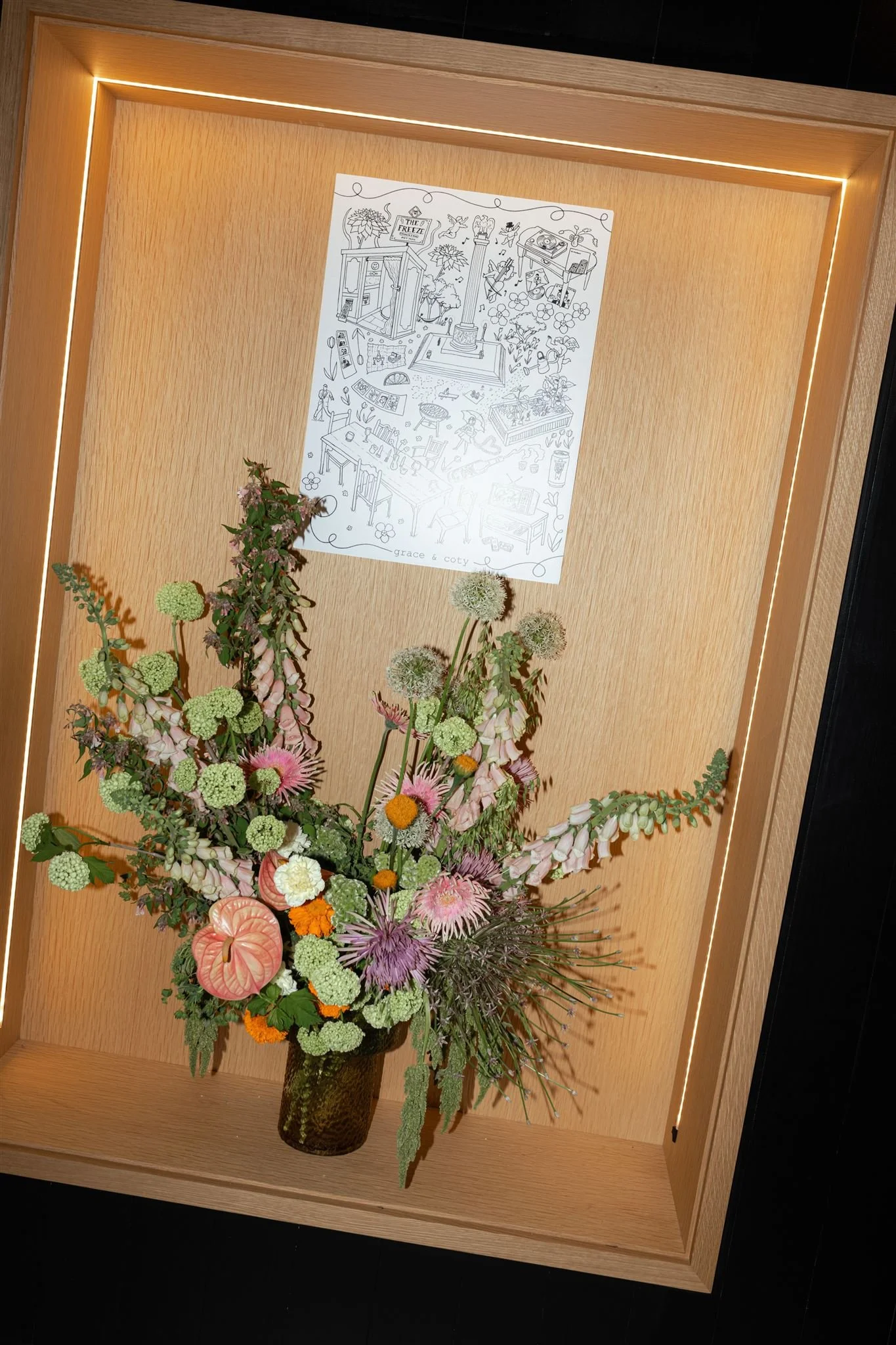 A floral arrangement with pink, white, green, and orange flowers in a dark vase, displayed on a wooden surface inside a wooden-framed display case with internal lighting, and a black-and-white line drawing poster on the back wall.