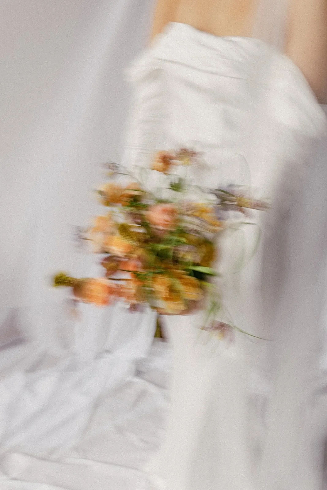 A blurry image of a person in white clothing holding a bouquet of orange and pink flowers against a plain background.