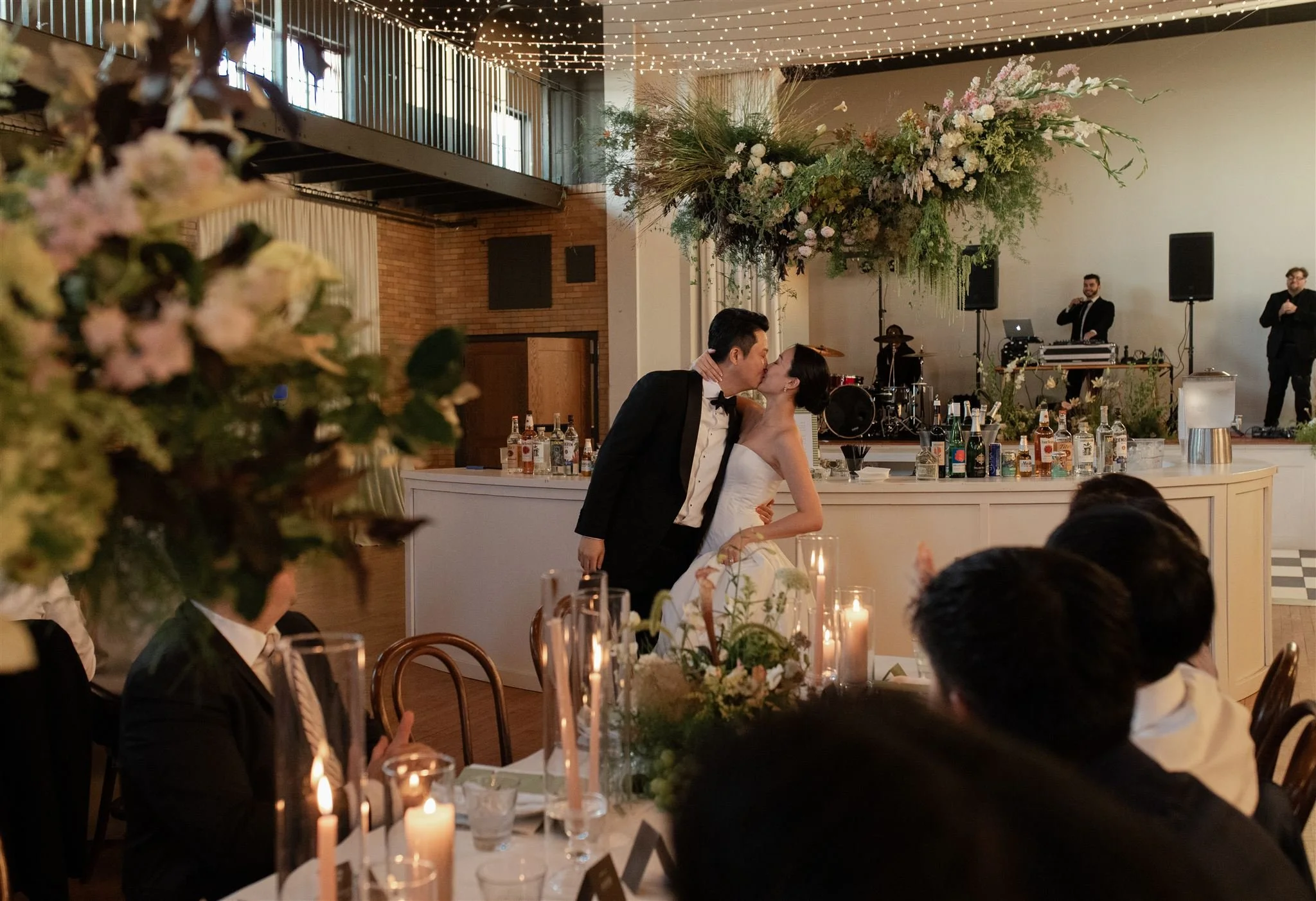 Bride and groom sharing a kiss at their wedding reception, surrounded by guests, candles, and floral arrangements, with a band playing on stage in the background.