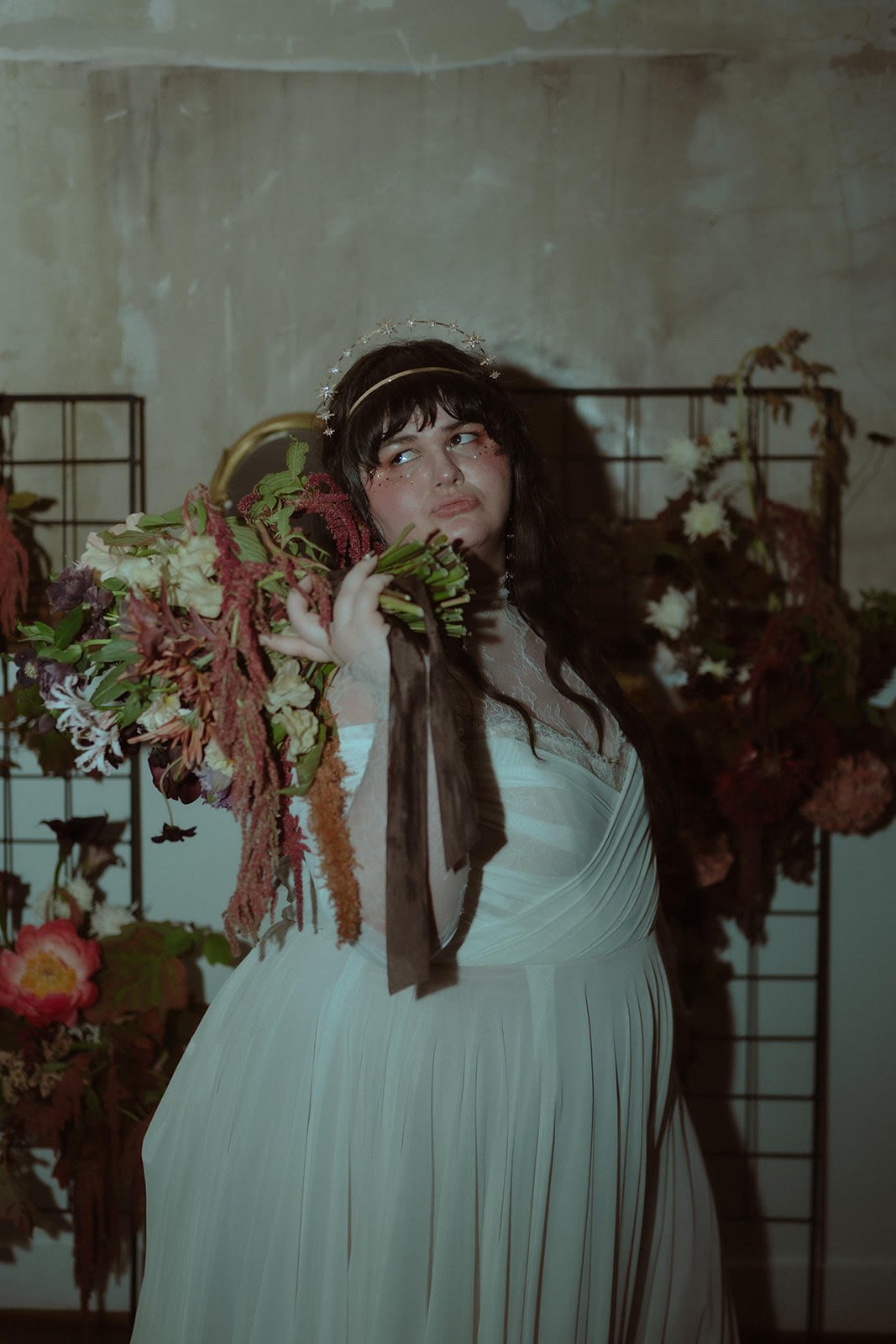 A woman dressed in a vintage white gown holding a bouquet of flowers in a dimly lit room with floral decorations and a metal grid background.