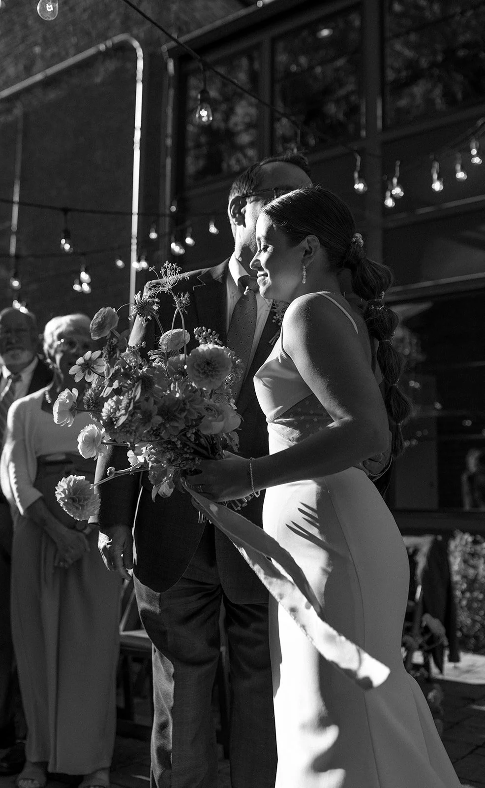 A black and white photo of a bride and groom sharing a moment during their wedding ceremony, standing close with the bride holding a bouquet of flowers, surrounded by guests under string lights.
