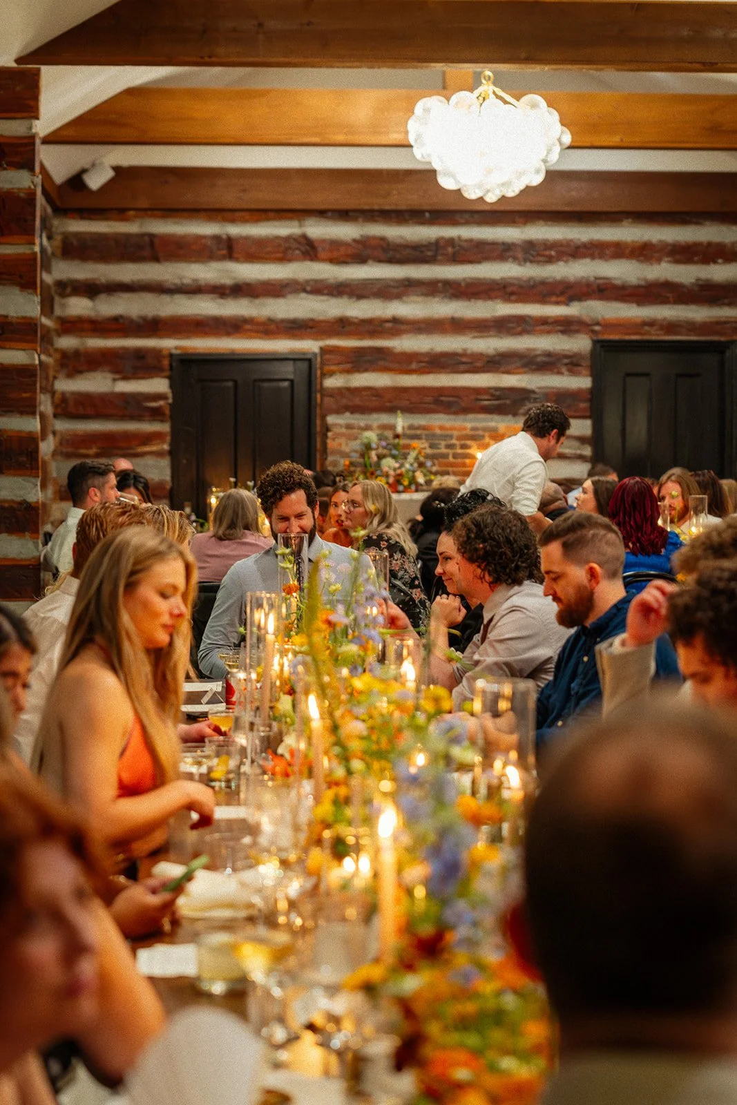 People dining at a long table decorated with flowers and candles in a rustic log cabin-style setting with wood-paneled walls and a large chandelier overhead.