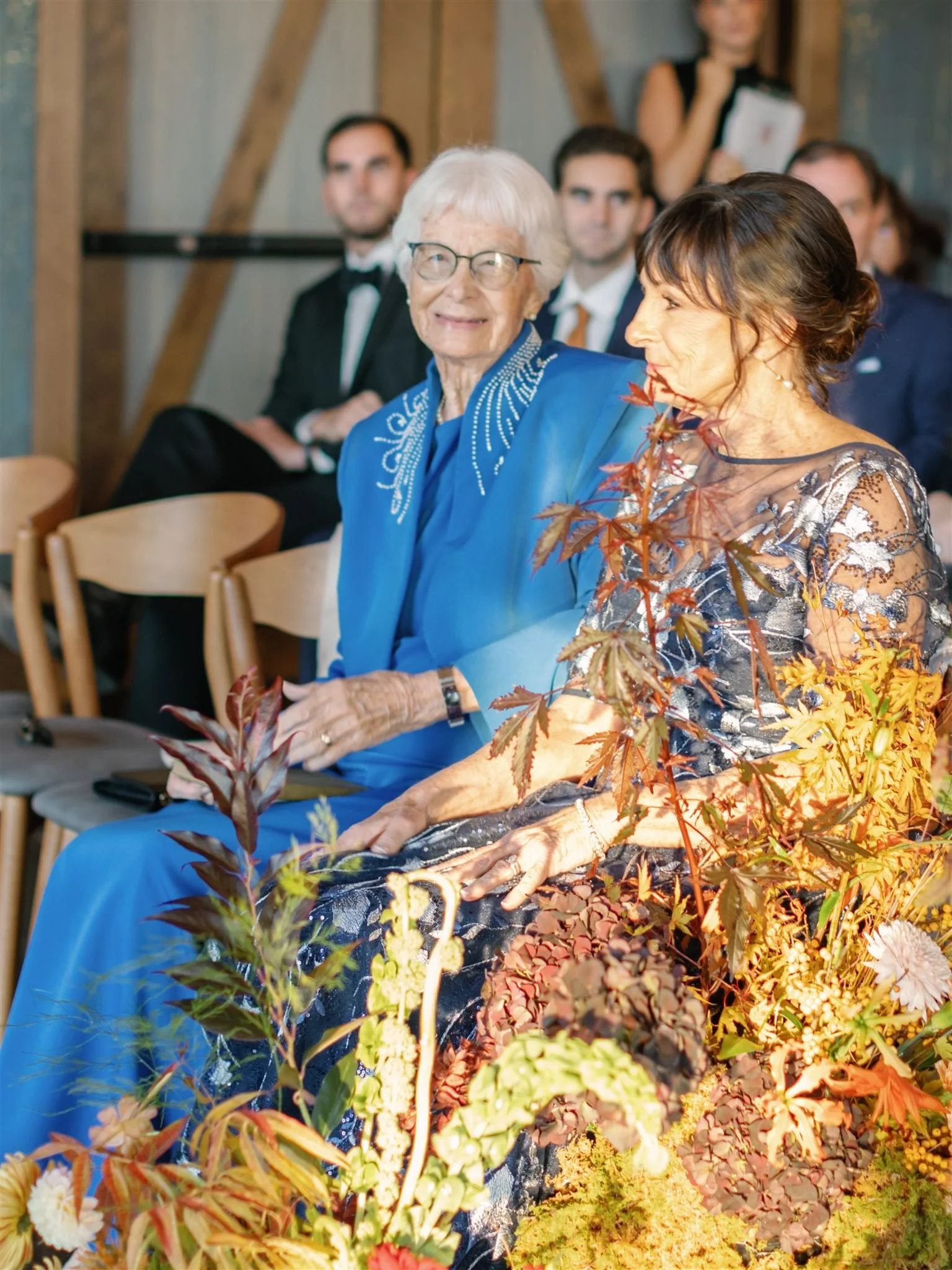 An elderly woman with white hair and glasses wearing a bright blue dress is sitting next to a woman in a floral dress. They are attending an indoor event with other people in formal attire in the background, and there are autumn-colored plants in the