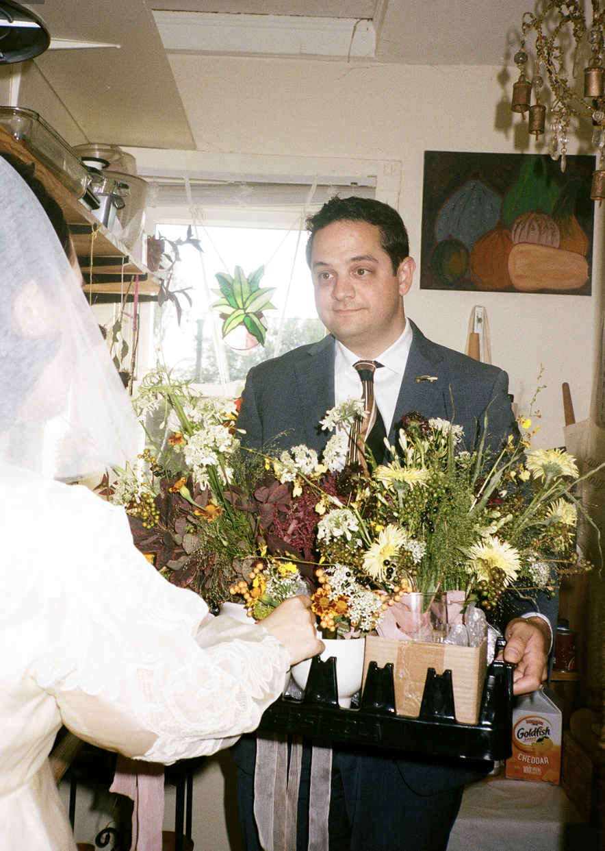 A man in a suit holding a tray with a large bouquet of flowers, standing in a room with artwork on the wall and a chandelier.