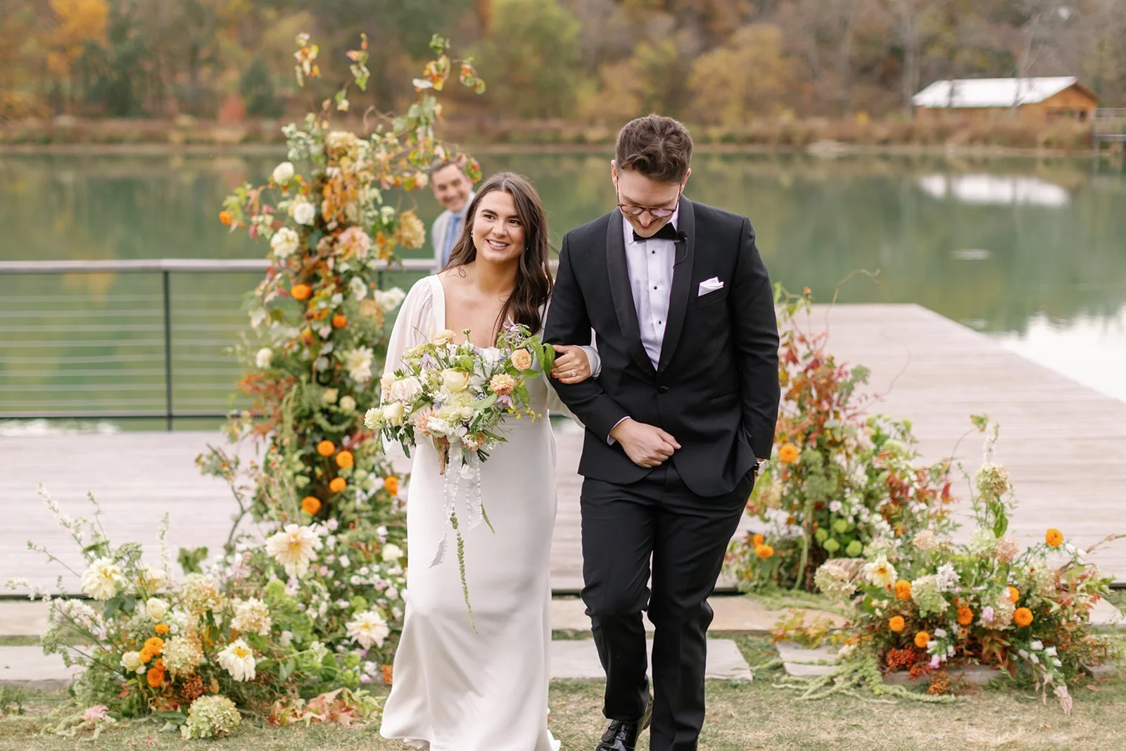 A bride and groom walking arm-in-arm down a path decorated with flowers at an outdoor wedding by a lake, with a groomsman in the background.