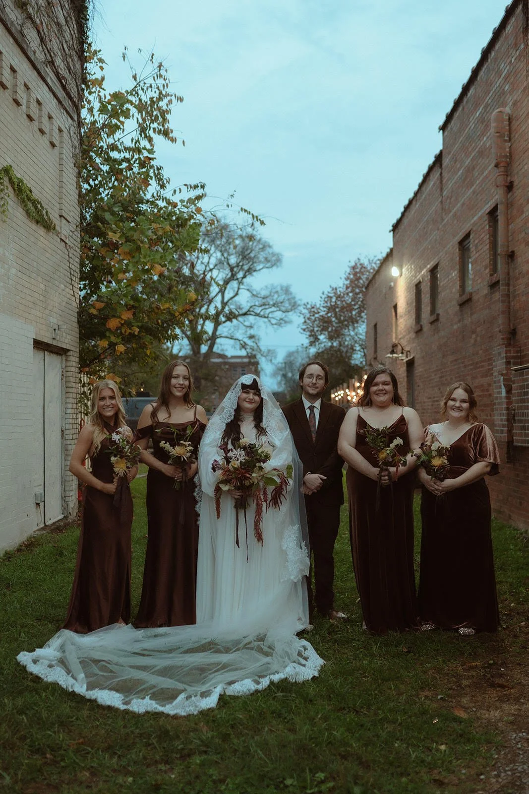 A wedding party standing outdoors between brick buildings in a narrow alley on a cloudy evening, with the bride and groom in the center, surrounded by bridesmaids holding bouquets, all smiling.