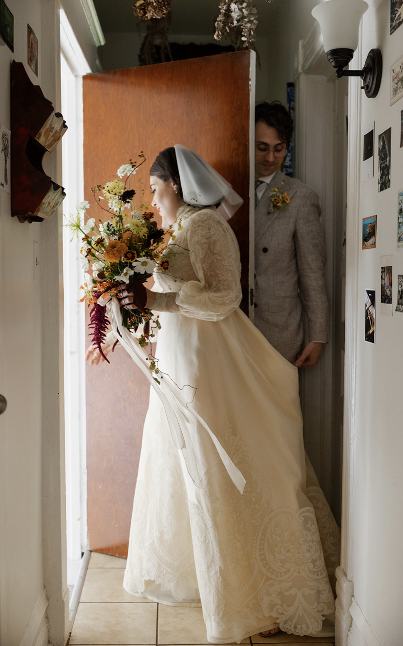 A bride and groom are peeking out from a doorway, with the bride holding a bouquet of flowers and looking down, wearing a vintage lace wedding dress and veil, and the groom in a gray suit with a boutonniere, in a cozy home interior decorated with wal