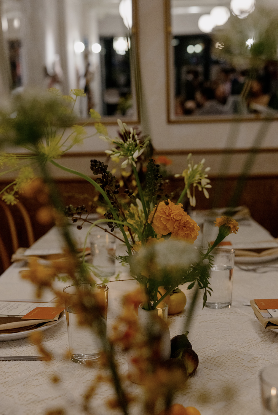 A close-up of a table centerpiece with a vase containing yellow and white flowers, with glasses, napkins, and place settings in the background at a dinner event.