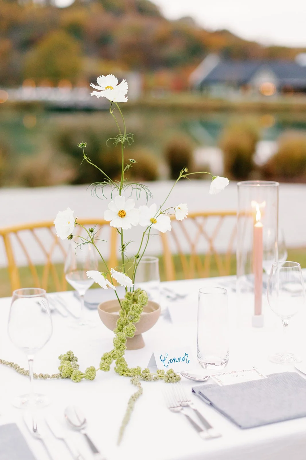 Elegant outdoor dining table with white tablecloth, floral centerpiece in a beige bowl, glassware, silverware, a pink candle in a glass holder, and a chair in the background, with a scenic autumn landscape.