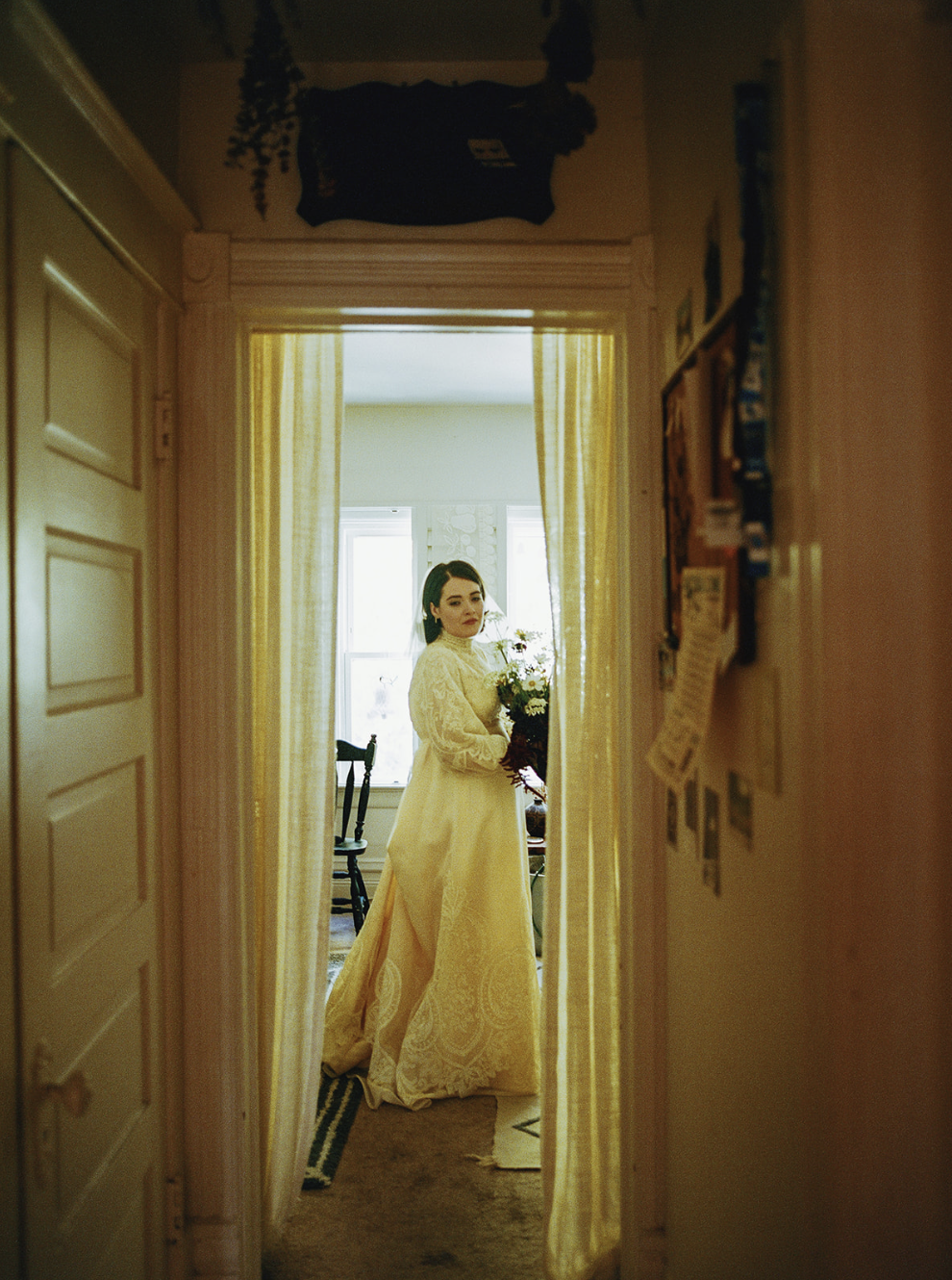 A woman in a vintage wedding dress holding a bouquet of flowers standing in a doorway in a cozy, sunlit room with yellow curtains.