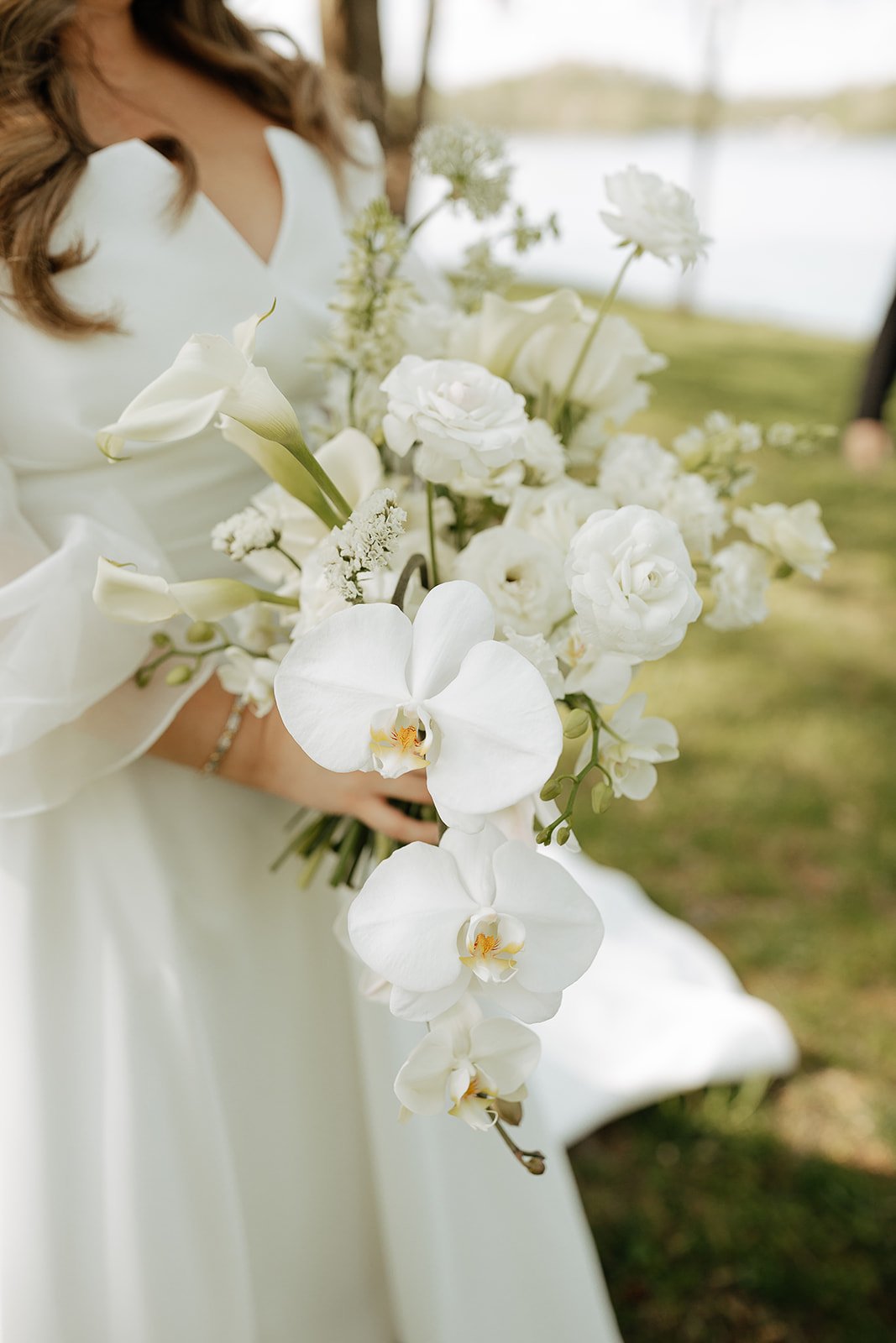 A woman in a white dress holding a large bouquet of white flowers, including orchids, roses, calla lilies, and other blooms, outdoors near a body of water.
