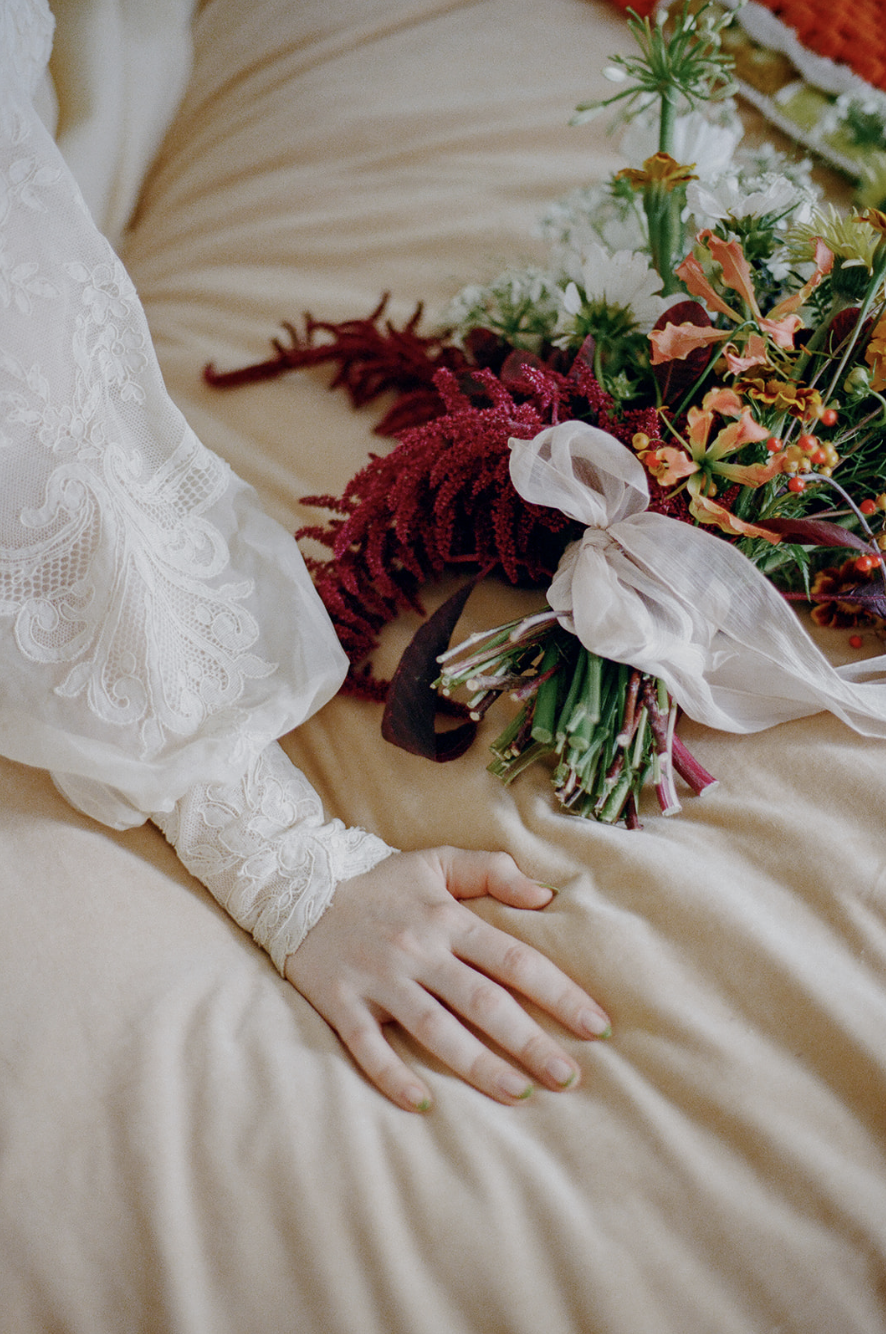 A person's hand resting on a beige bedspread near a bouquet of colorful flowers tied with a ribbon.